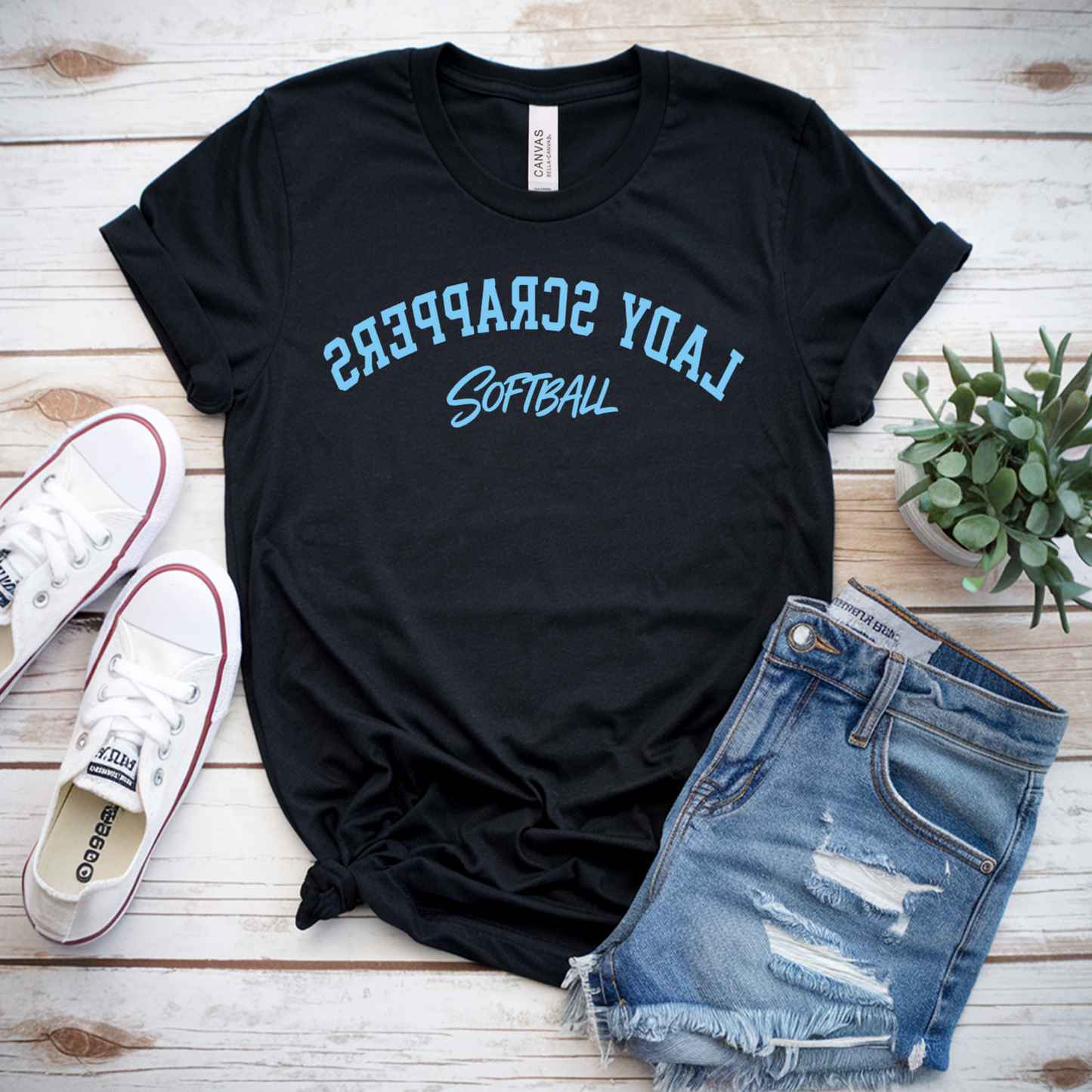 Black t-shirt with 'Lady Scorpions Softball' text, white sneakers, and denim shorts on a wooden surface.