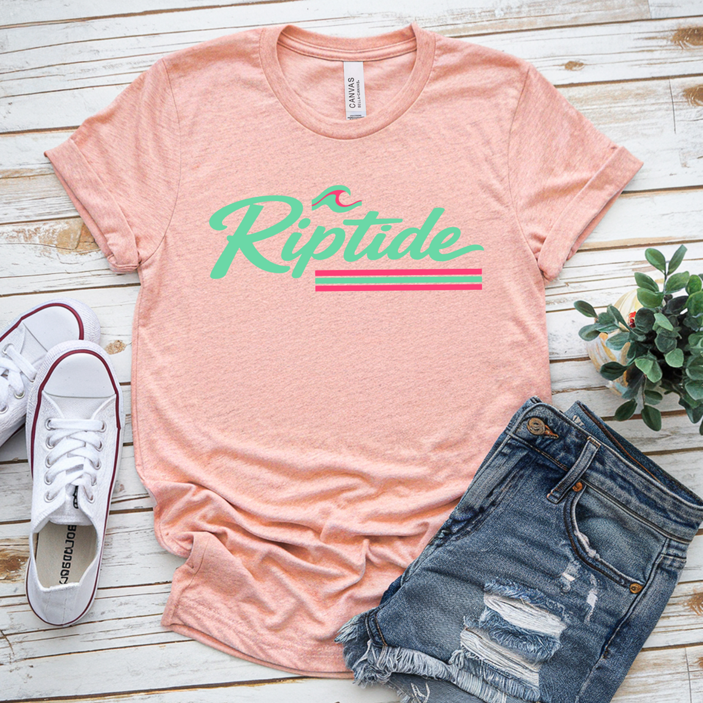Pink t-shirt with 'Riptide' design, white sneakers, and denim shorts on a wooden surface.