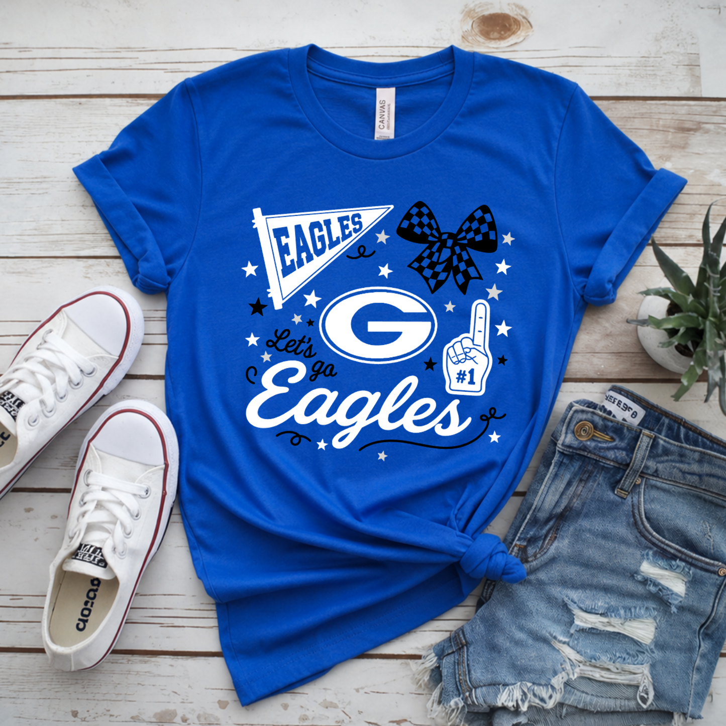 A royal blue graphic T-shirt laid flat on a whitewashed wood surface, styled with white sneakers, distressed denim shorts, and a small potted plant. The shirt graphic reads "Eagles" with a large "G" logo, stars, a bow tie, and a hand gesture.