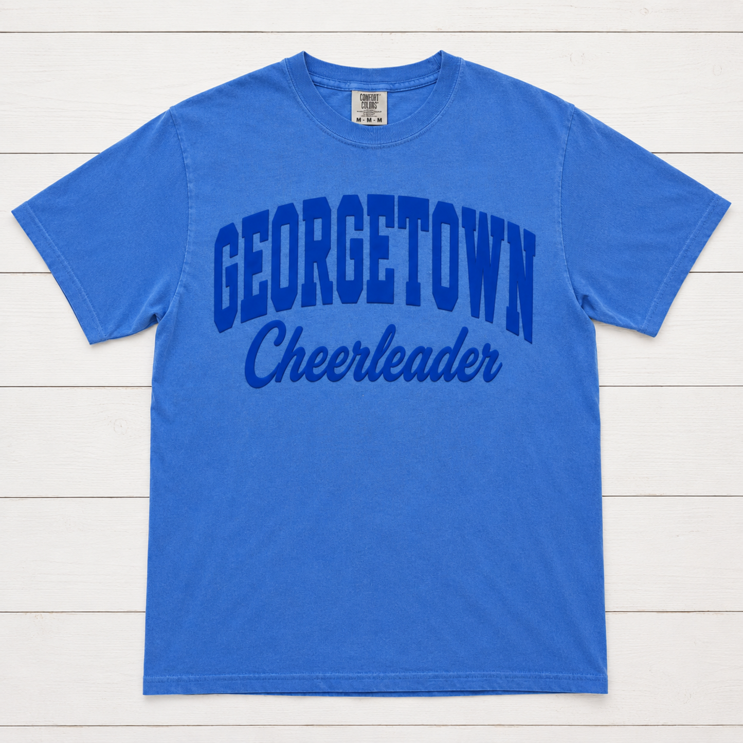 A royal blue short-sleeve t-shirt with 'GEORGETOWN Cheerleader' printed in dark blue across the front, laid flat on a white wood plank background.