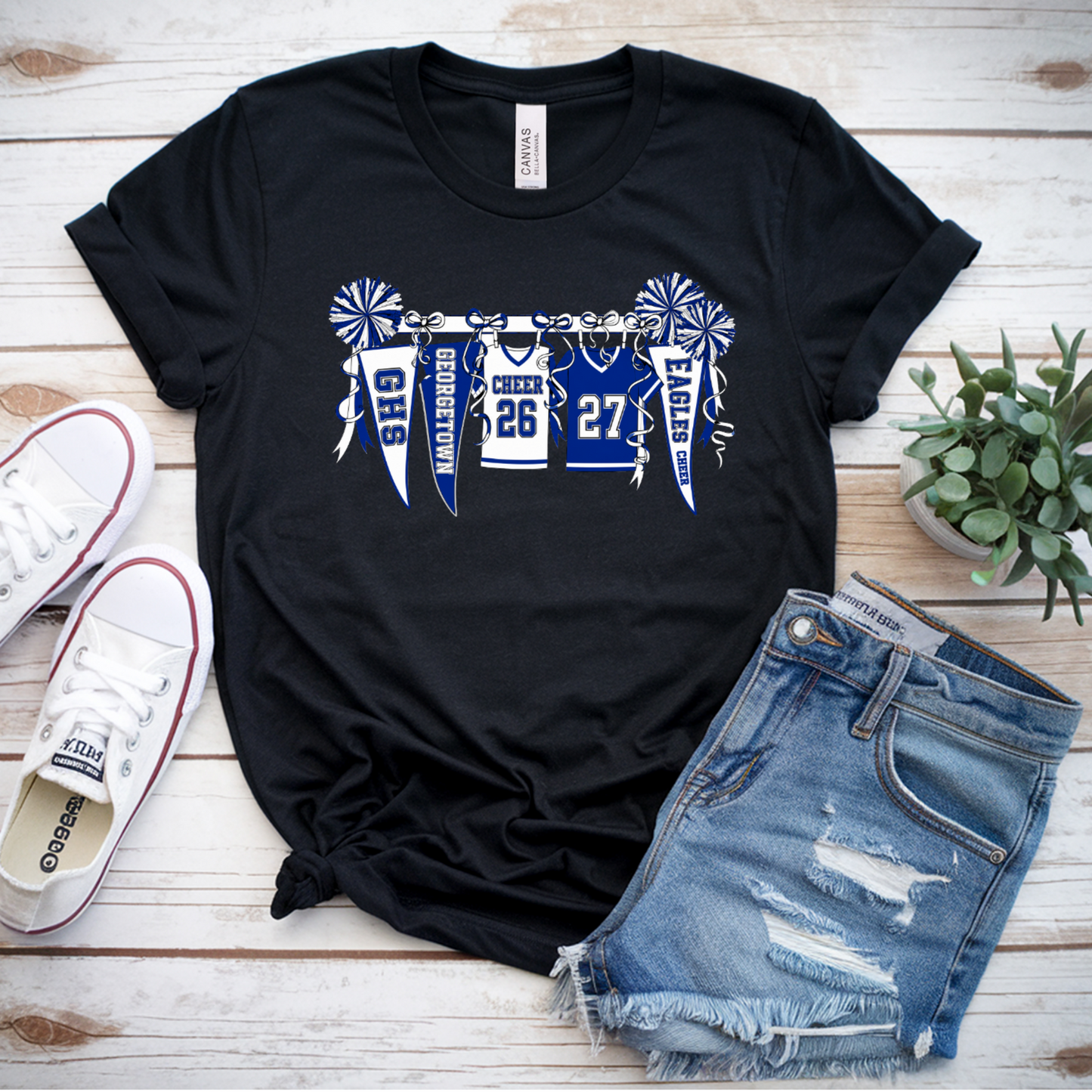 A black graphic t-shirt featuring a blue and white 'Buckeyes 2024' football jersey design, styled in a flat lay with white sneakers, denim shorts, and a small potted succulent on a light wood background.