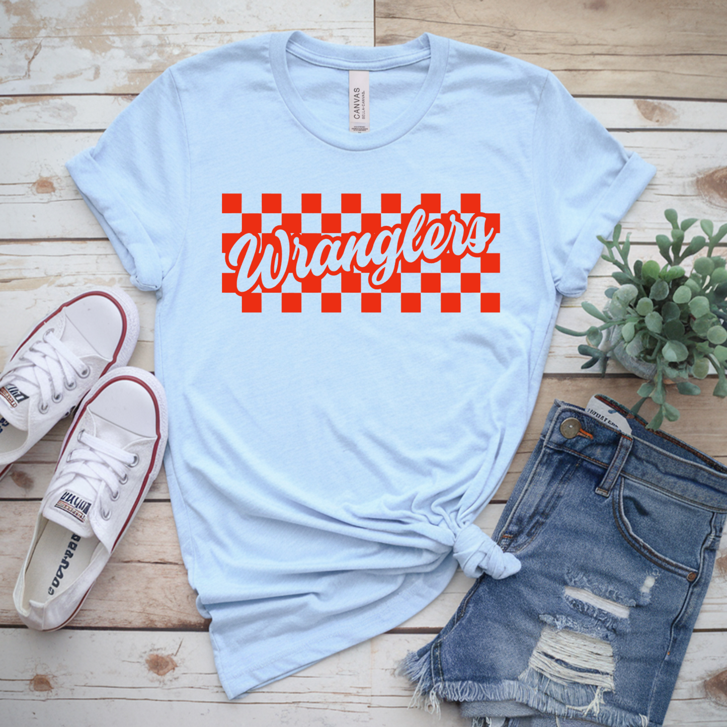 Light blue t-shirt with red and white checkered 'Wrangler' logo on a wooden surface with sneakers and shorts.