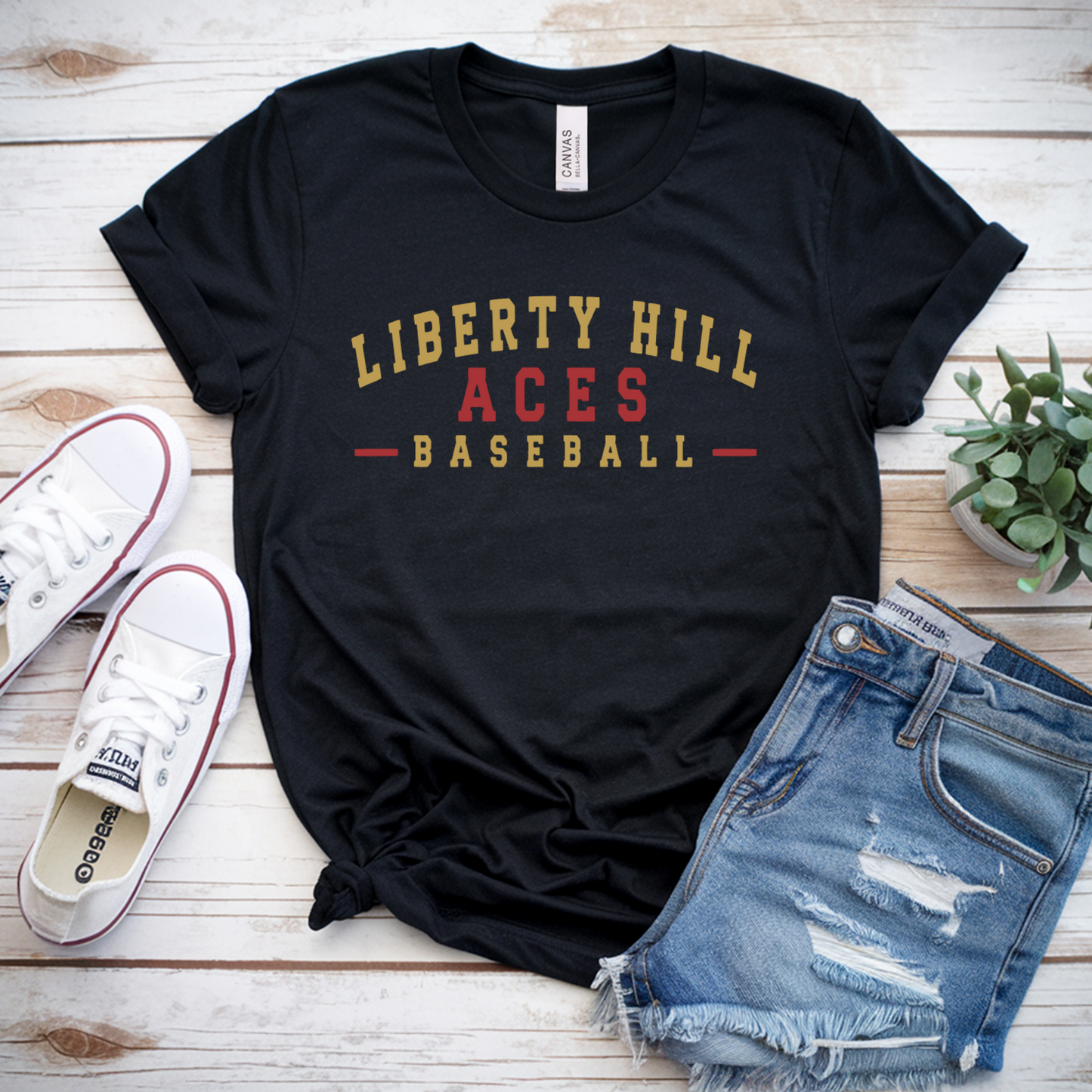 A black graphic t-shirt with 'LIBERTY HILL ACES BASEBALL' printed in gold and red, styled in a flat lay with blue denim shorts, white Converse sneakers, and a small potted succulent on a white wood plank background.