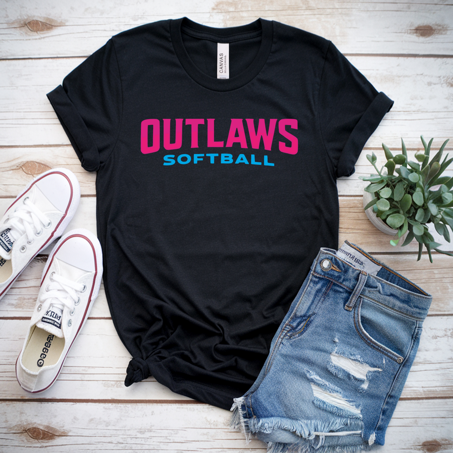 Black t-shirt with 'OUTLAWS SOFTBALL' text, white sneakers, and denim shorts on a wooden surface.