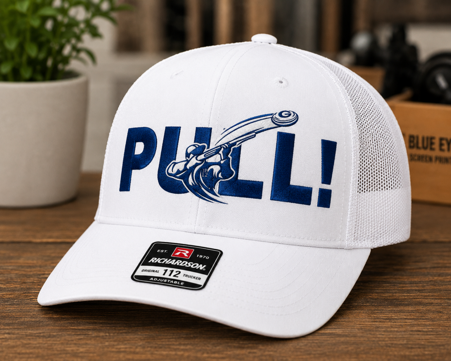 A white Richardson 112 trucker cap with a blue 'PULL!' graphic on the front, displayed on a wooden surface with a blurred indoor background.