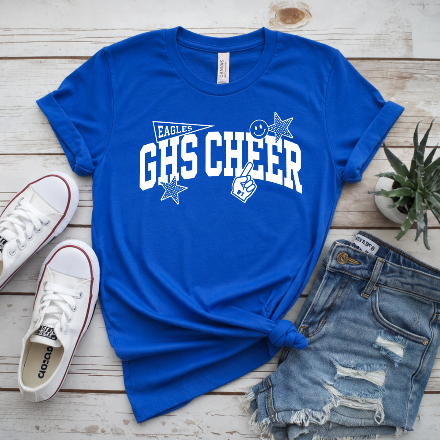 A royal blue graphic T-shirt with distressed denim shorts, white sneakers, and a small potted succulent arranged on a whitewashed wood surface. The shirt features the text 'EAGLES GHS CHEER' in white with star and ribbon graphics.