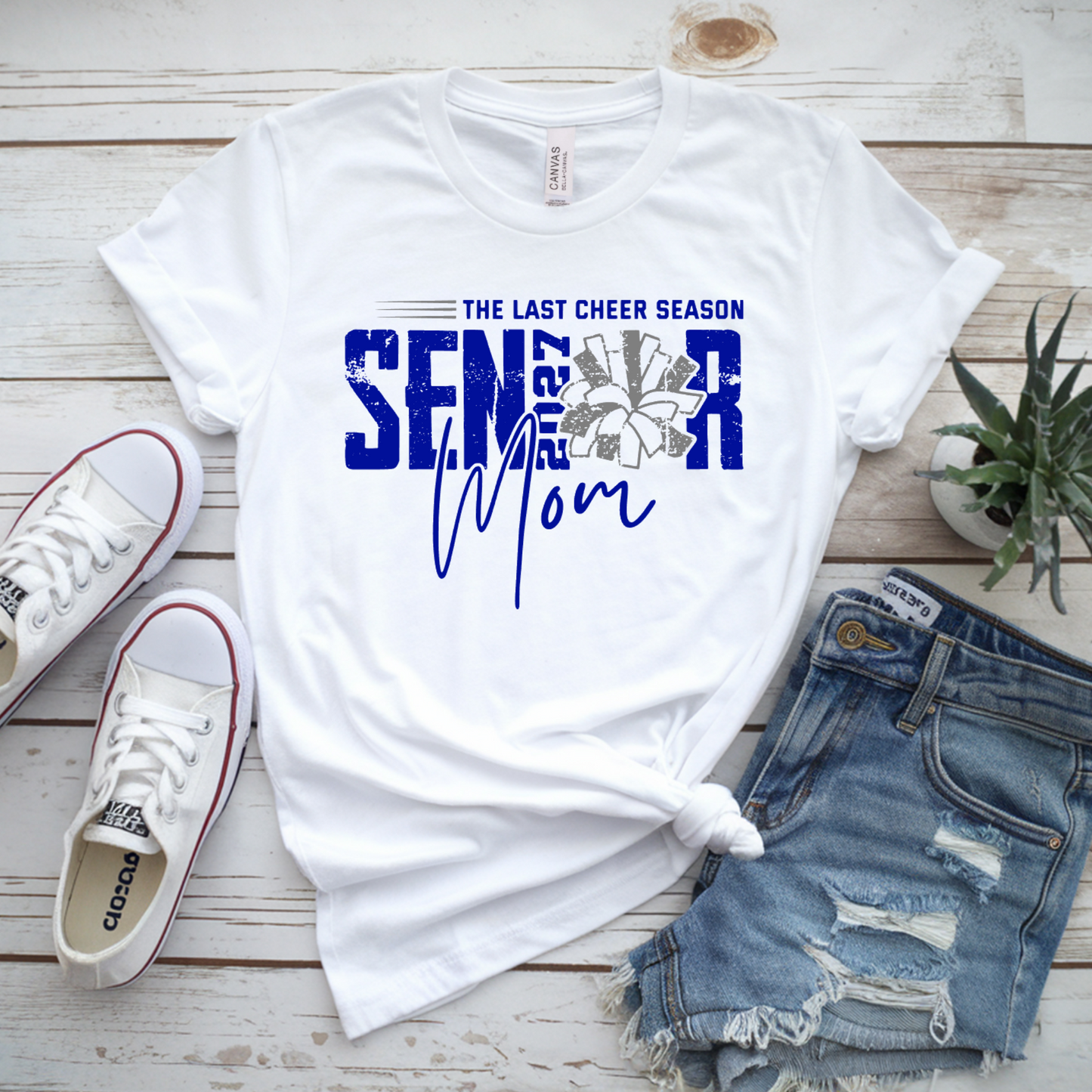 White graphic T-shirt laid flat on a whitewashed wood surface, styled with white sneakers, distressed denim shorts, and a small potted plant. The shirt reads 'THE LAST CHEER SEASON SENIOR Mom' in blue and gray text with a floral design.