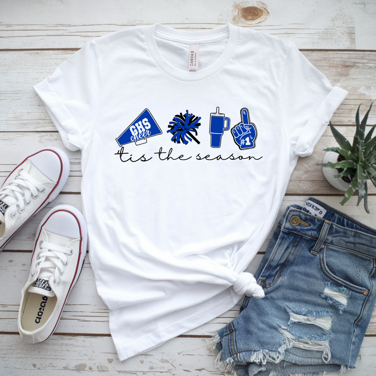 White graphic T-shirt laid flat on a whitewashed wood surface, styled with white Converse sneakers, distressed denim shorts, and a small potted succulent. The shirt has a blue cheer-themed graphic and the words 'tis the season' printed on the front.