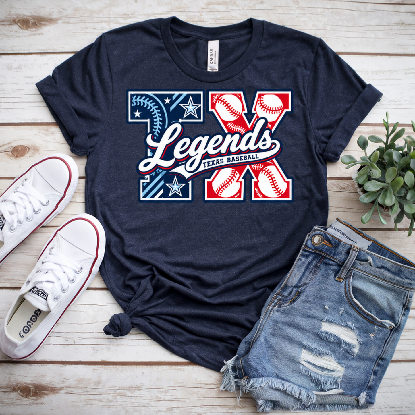 A navy blue graphic T-shirt styled in a flat lay on a light wood background, paired with white sneakers, distressed denim shorts, and a small potted succulent. The shirt features a large 'XX Legends Texas Baseball' graphic in red, white, and blue.