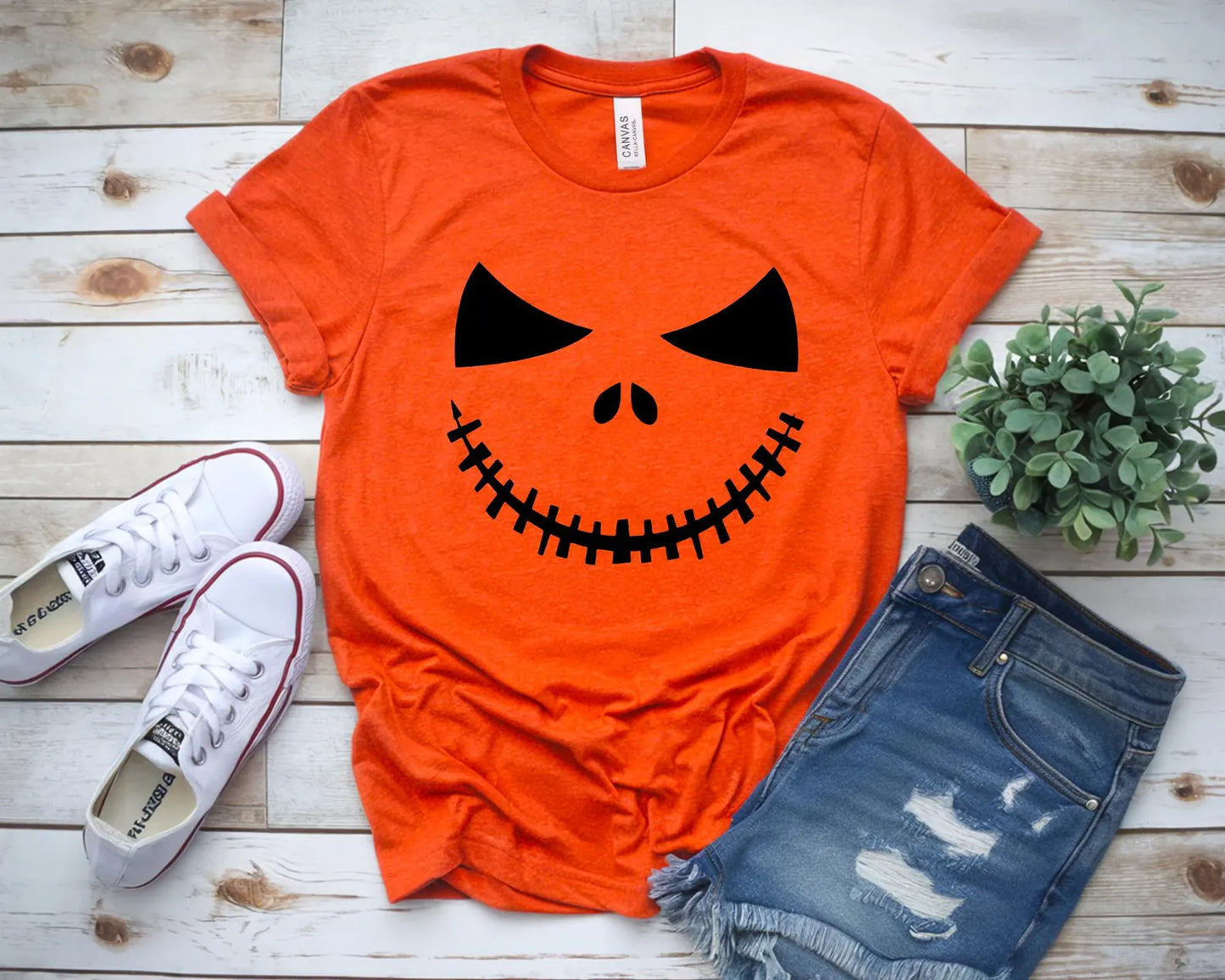 Orange t-shirt with a pumpkin face design, paired with white sneakers and denim shorts on a wooden surface.