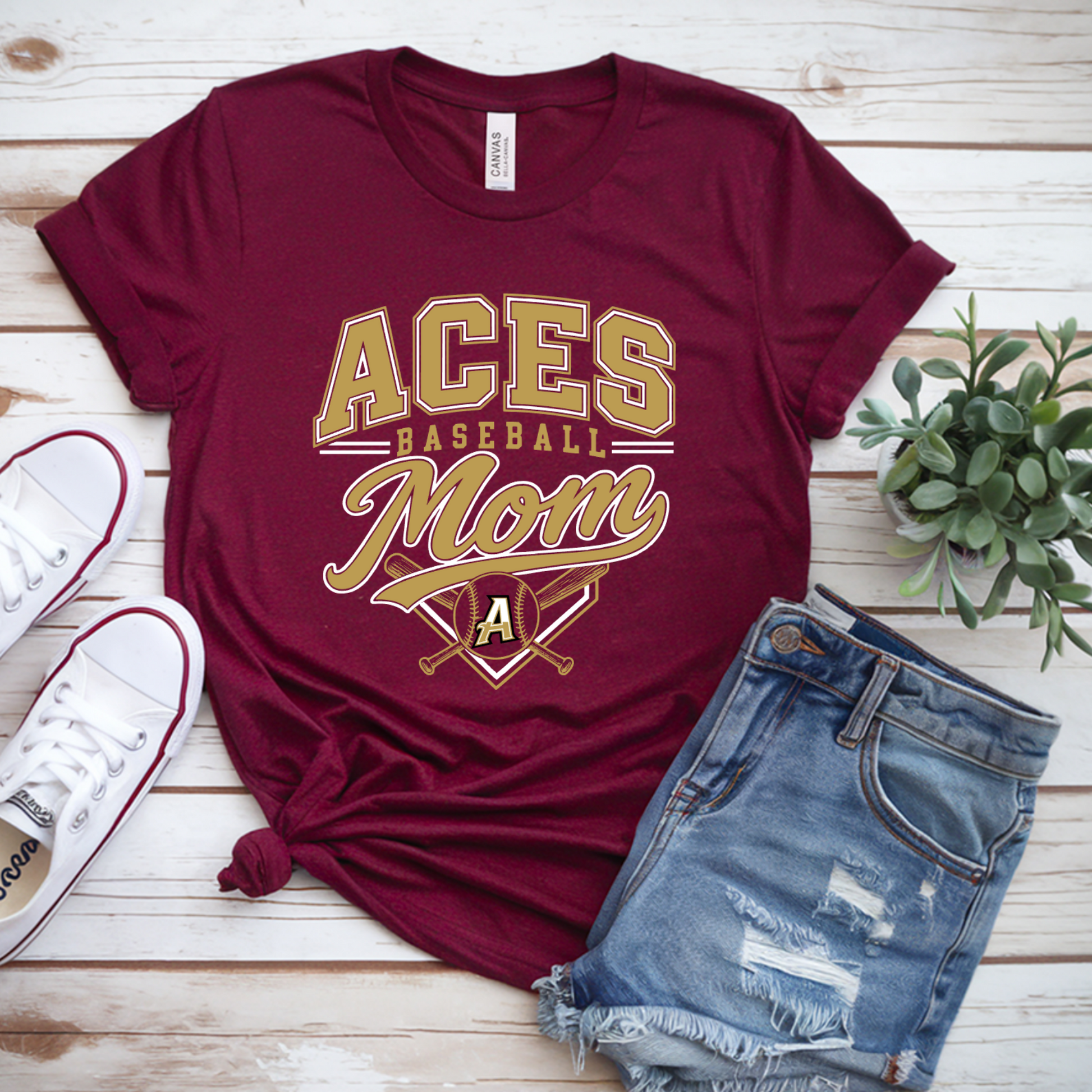 A maroon short-sleeve graphic T-shirt laid flat on a whitewashed wood surface, styled with white sneakers, distressed denim shorts, and a small green plant. The shirt reads "ACES BASEBALL Mom" in gold and white lettering with crossed bats and a letter A graphic.