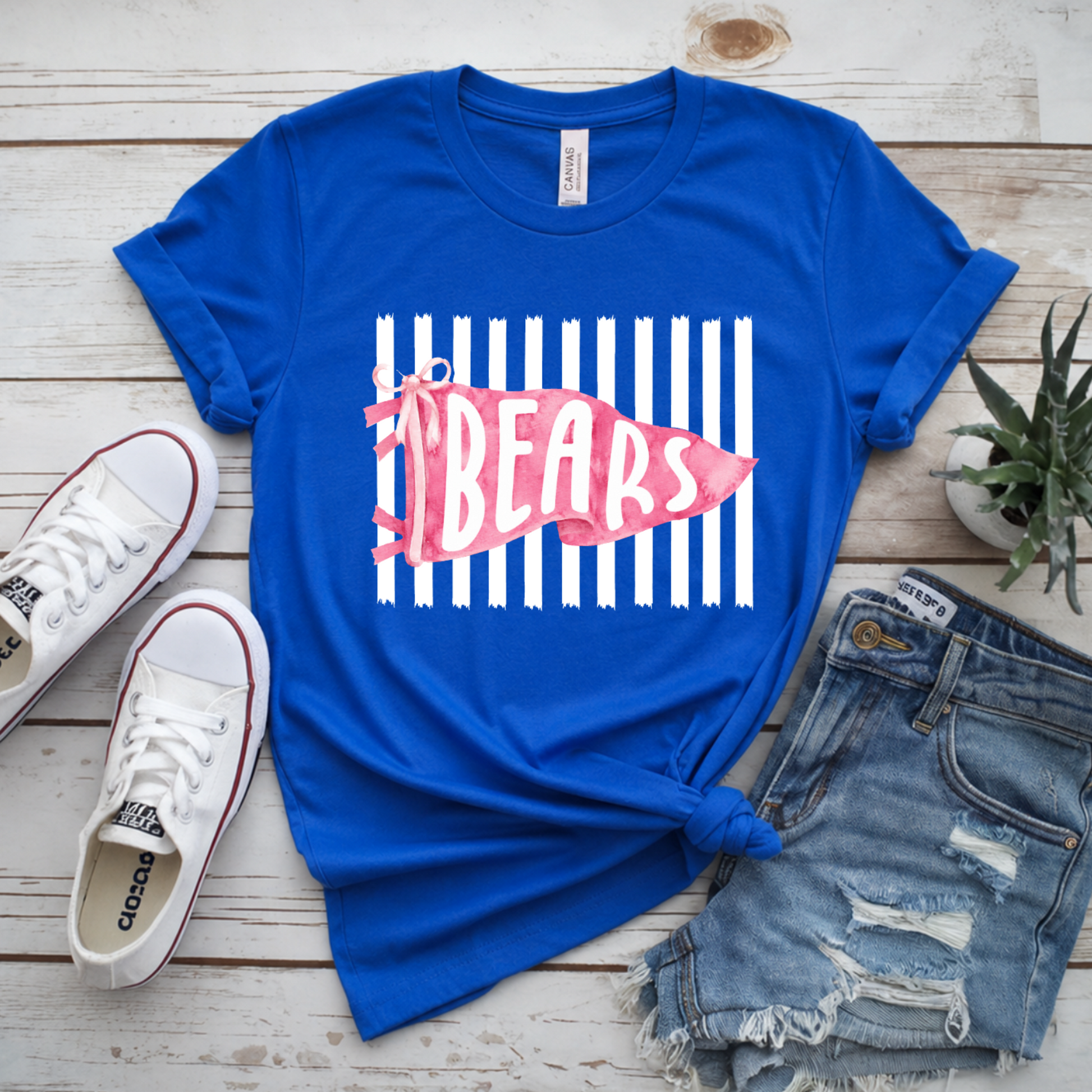 A royal blue graphic T-shirt laid flat on a whitewashed wood surface, styled with white sneakers, distressed denim shorts, and a small potted plant. The shirt has a white vertical stripe graphic with a pink fish-shaped logo reading 'BEARS'.