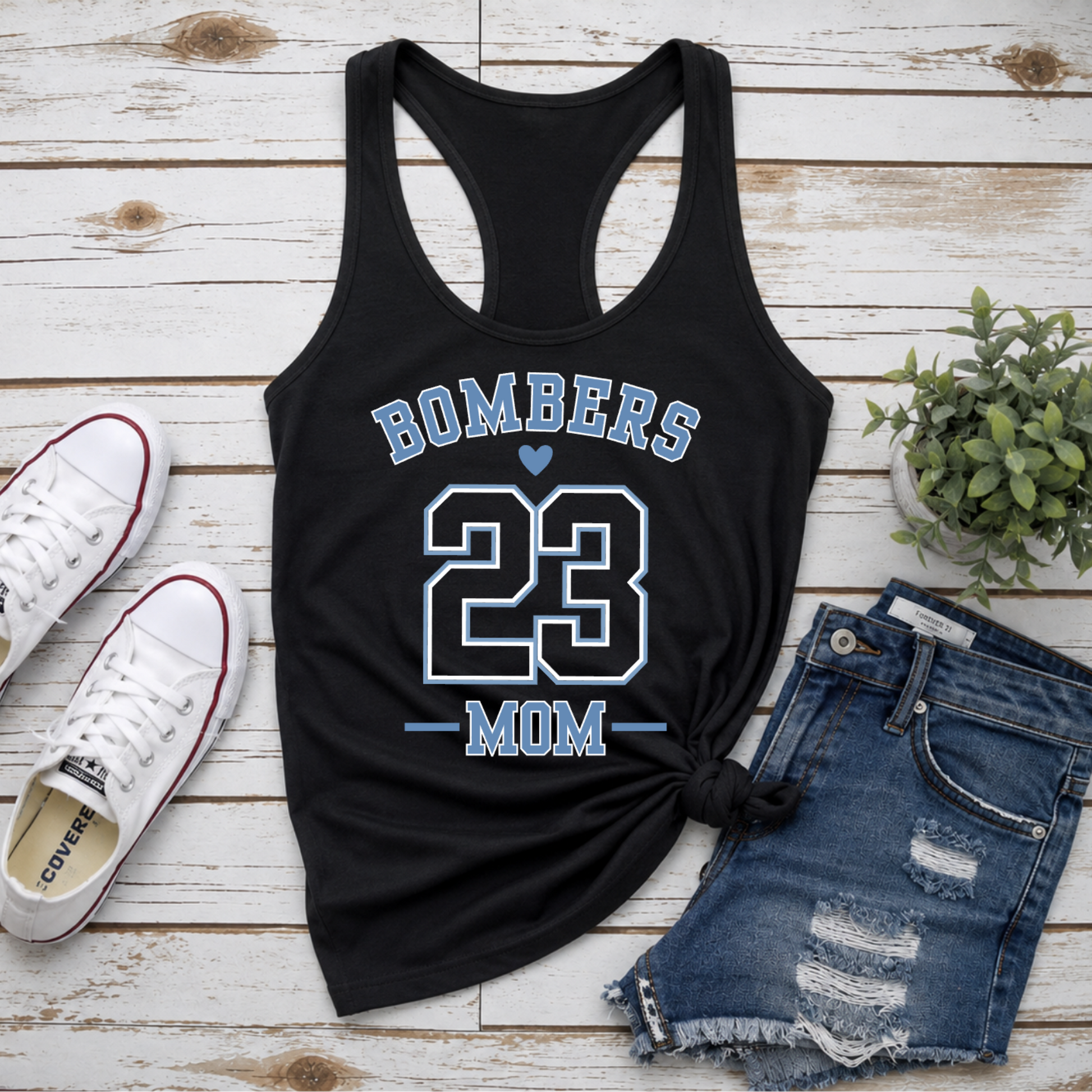 Black tank top with 'Bombers 23 Mom' text, white sneakers, denim shorts, and a plant on a wooden surface.