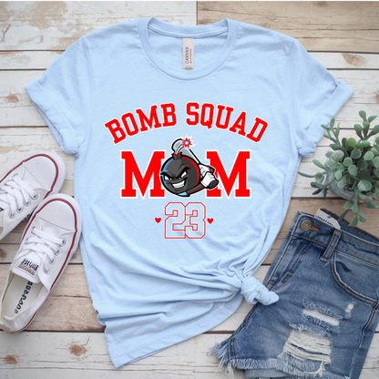 A light blue graphic T-shirt laid flat on a rustic wood surface, styled with white sneakers, distressed denim shorts, and a small green plant. The shirt reads 'BOMB SQUAD MOM 23' in red and black with a football helmet graphic.