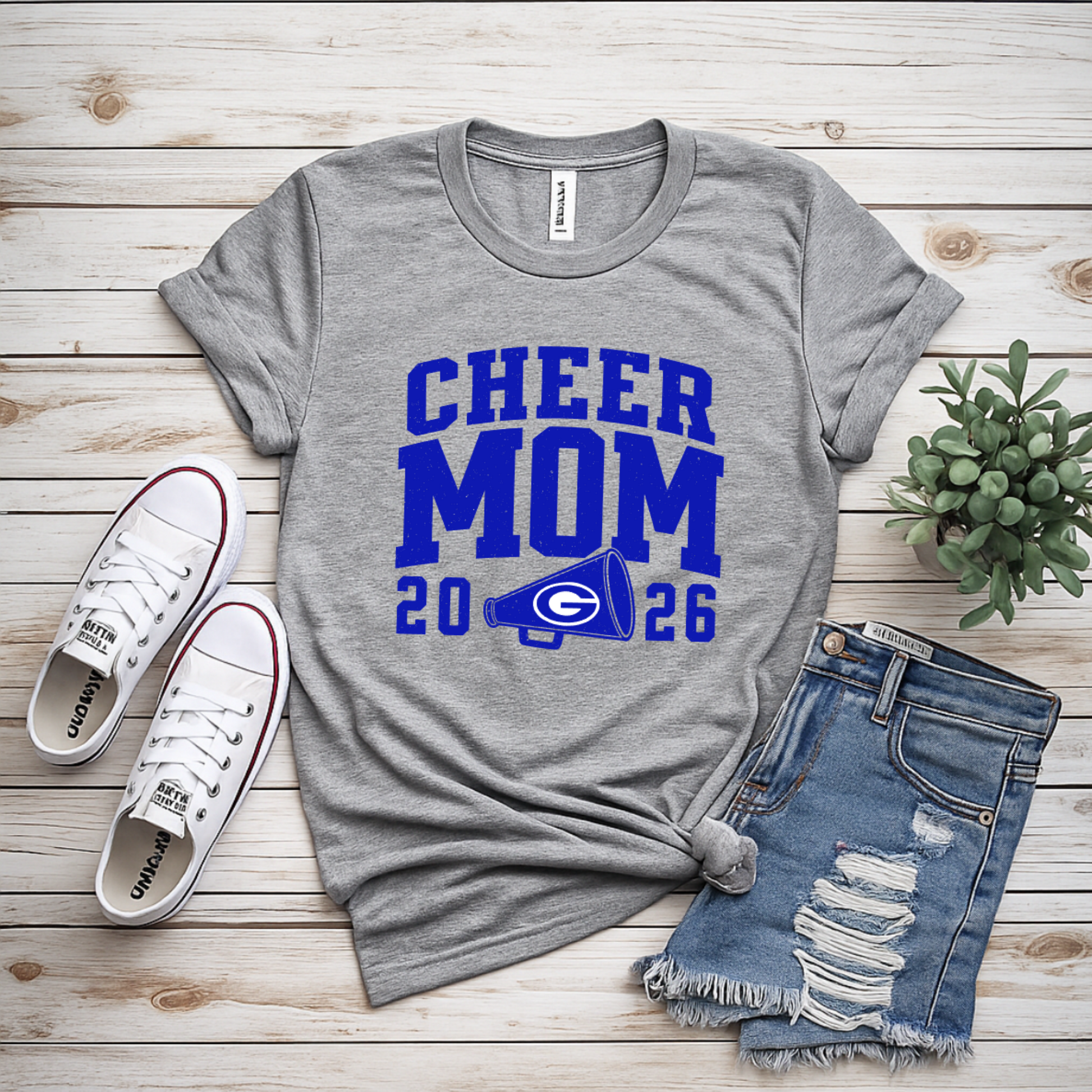 A gray heather graphic T-shirt with blue 'CHEER MOM 2026' text and a football logo, styled in a flat lay on a light wood plank background with white sneakers, distressed denim shorts, and a small succulent.