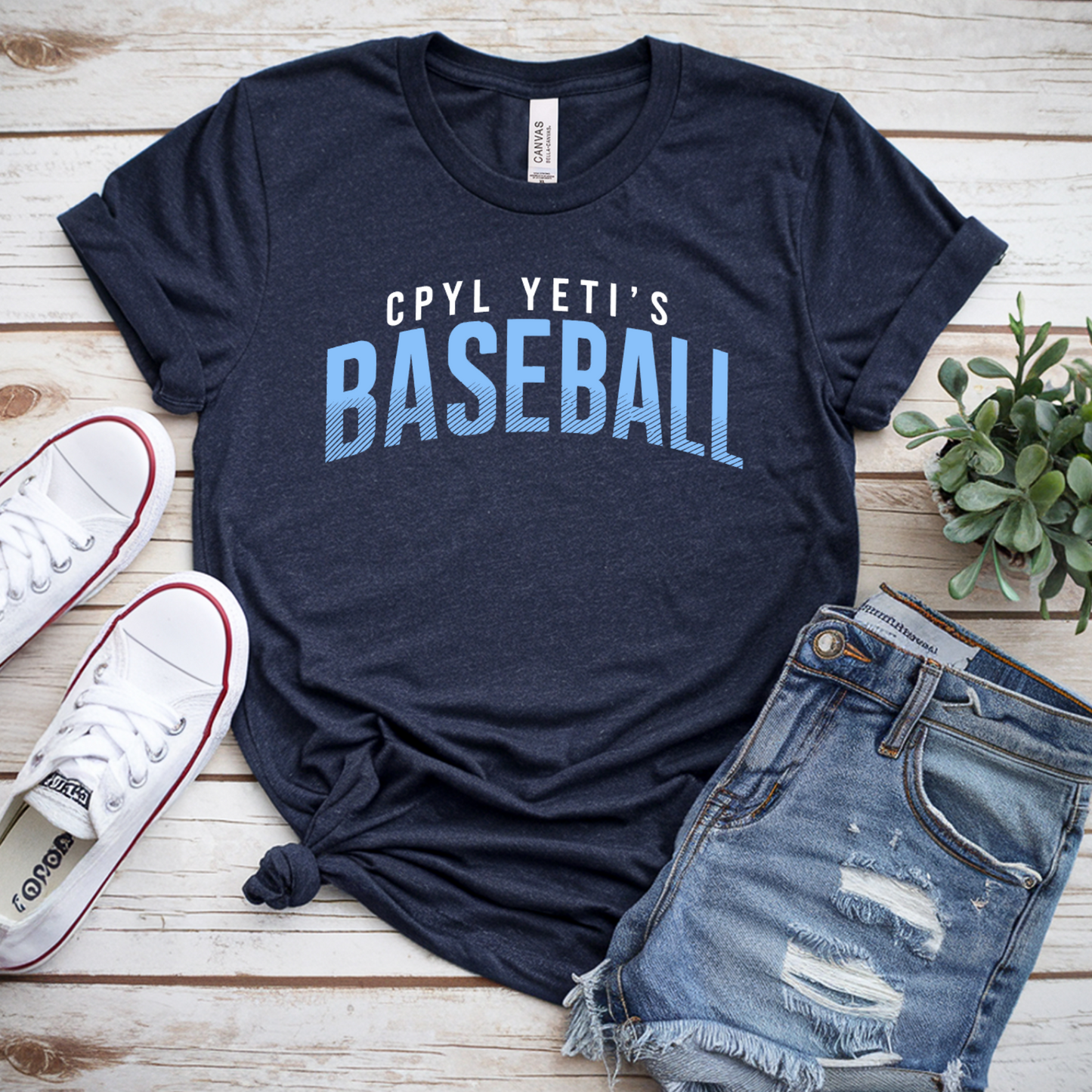 Navy t-shirt with 'CPYL YETI'S BASEBALL' text, white sneakers, denim shorts, and a plant on a wooden surface.