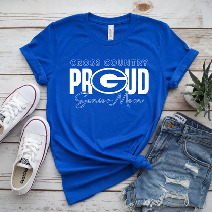 A bright blue short-sleeve T-shirt with a white 'Cross Country Proud Senior Mom' graphic, styled in a flat lay on a light wood background with white sneakers, distressed denim shorts, and a small potted succulent.