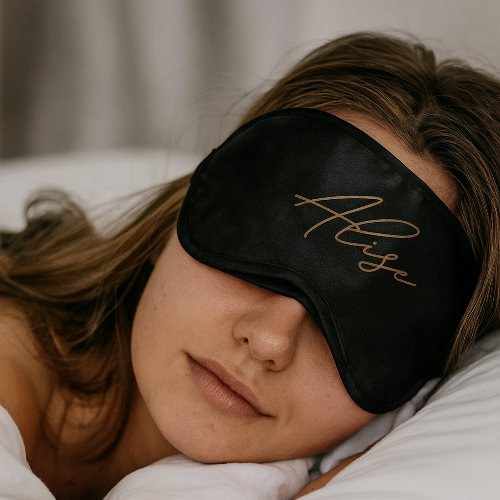 Person wearing a black sleep mask with 'Aliss' embroidered on it, lying in bed.