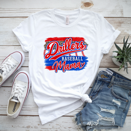 White graphic T-shirt laid flat on a whitewashed wood surface, styled with white sneakers, distressed denim shorts, and a small potted succulent. The shirt has a large red, white, and blue 'Drillers Baseball Mama' graphic on the front.