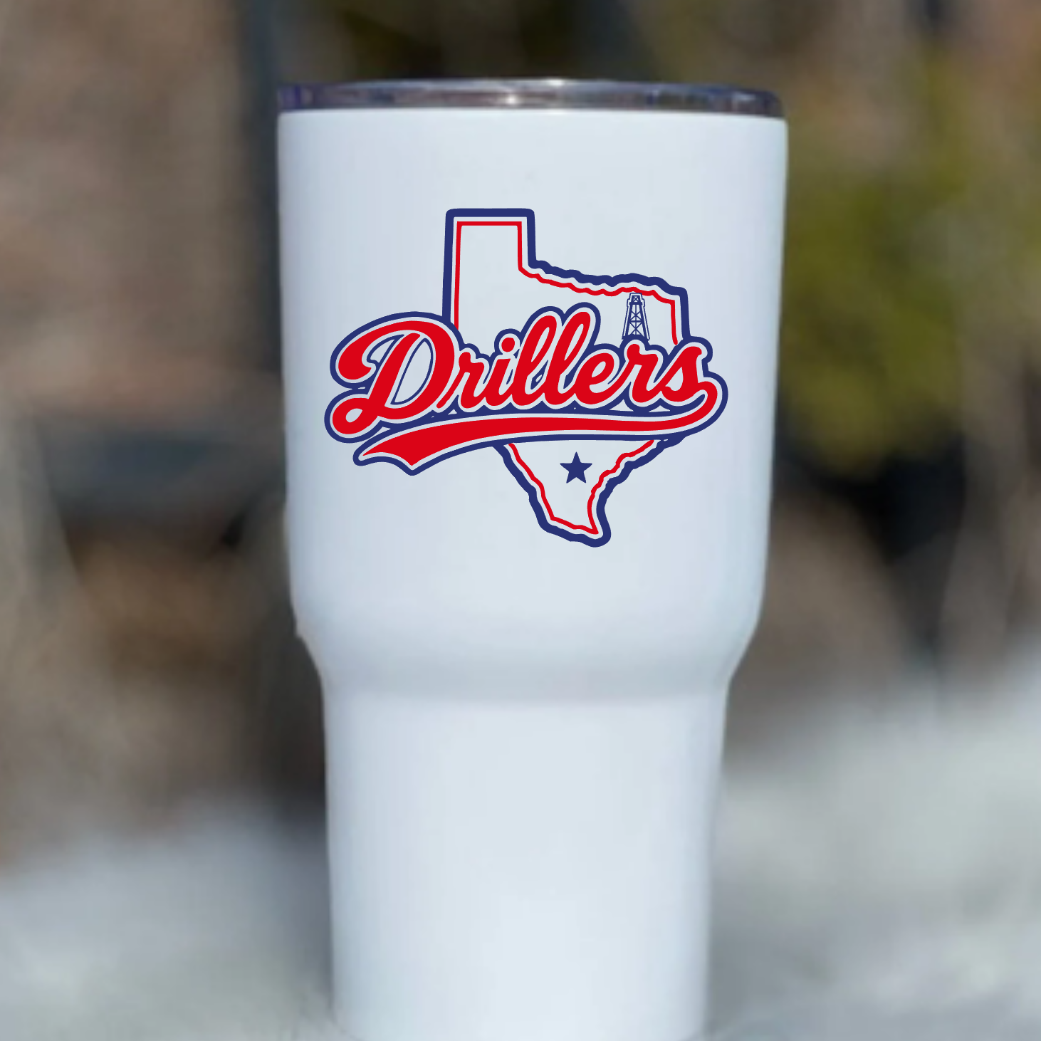 A white insulated tumbler with a clear lid, featuring a red and blue 'Drillers' logo over a Texas-shaped outline on the front, photographed against a softly blurred outdoor background.