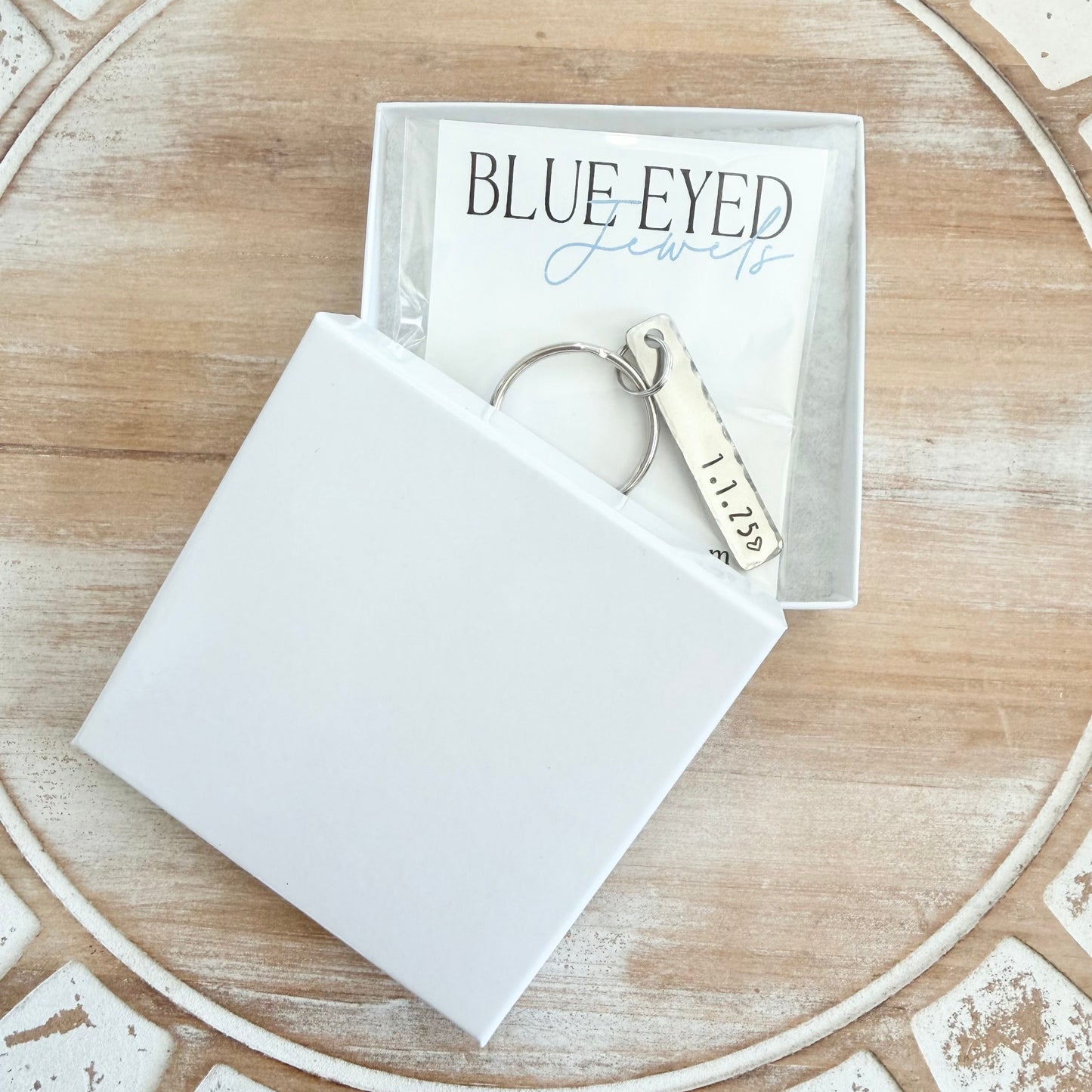 White jewelry box with a keychain and 'Blue Eyed' packaging on a decorative surface.