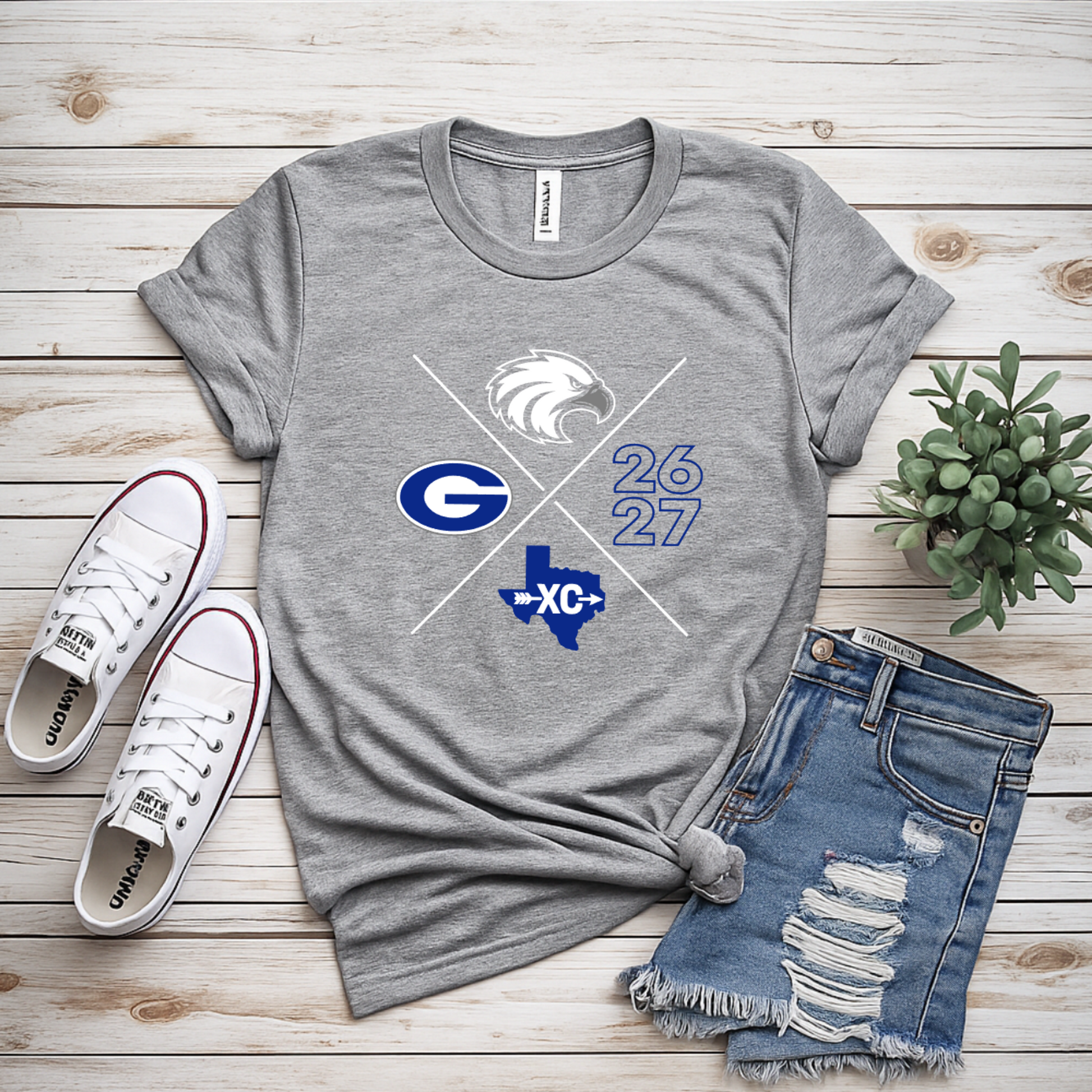 A heather gray graphic T-shirt laid flat on a light wood plank surface, styled with white sneakers, distressed denim shorts, and a small potted succulent. The shirt graphic shows a white eagle head, crossed white and blue diagonal lines, a blue 'G', the numbers '26 27', and a blue Texas outline with 'XC' inside.