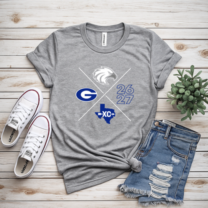 A heather gray graphic T-shirt laid flat on a light wood plank surface, styled with white sneakers, distressed denim shorts, and a small potted succulent. The shirt graphic shows a white eagle head, crossed white and blue diagonal lines, a blue 'G', the numbers '26 27', and a blue Texas outline with 'XC' inside.