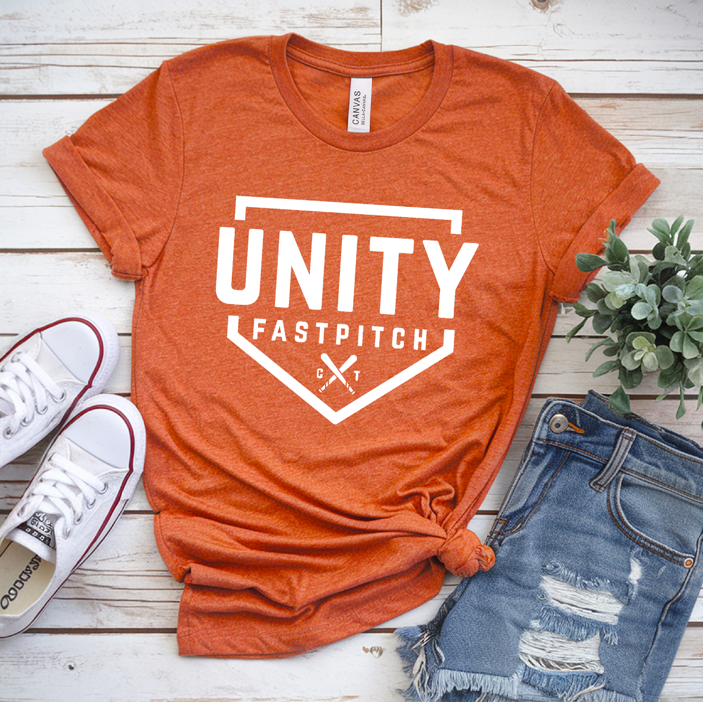 Orange t-shirt with 'Unity Fastpitch' design, white sneakers, and denim shorts on a wooden surface.