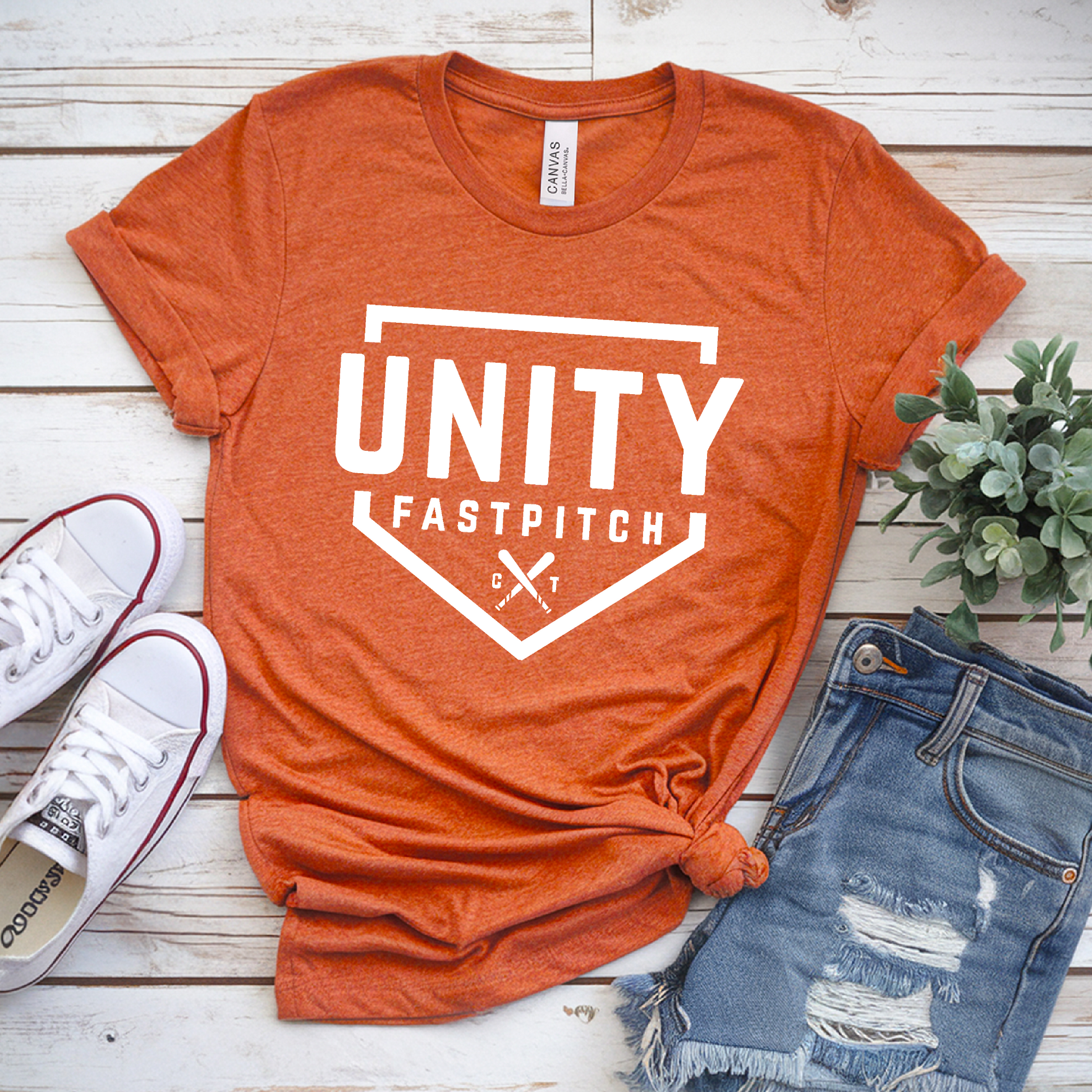 Orange t-shirt with 'Unity Fastpitch' design, white sneakers, and denim shorts on a wooden surface.