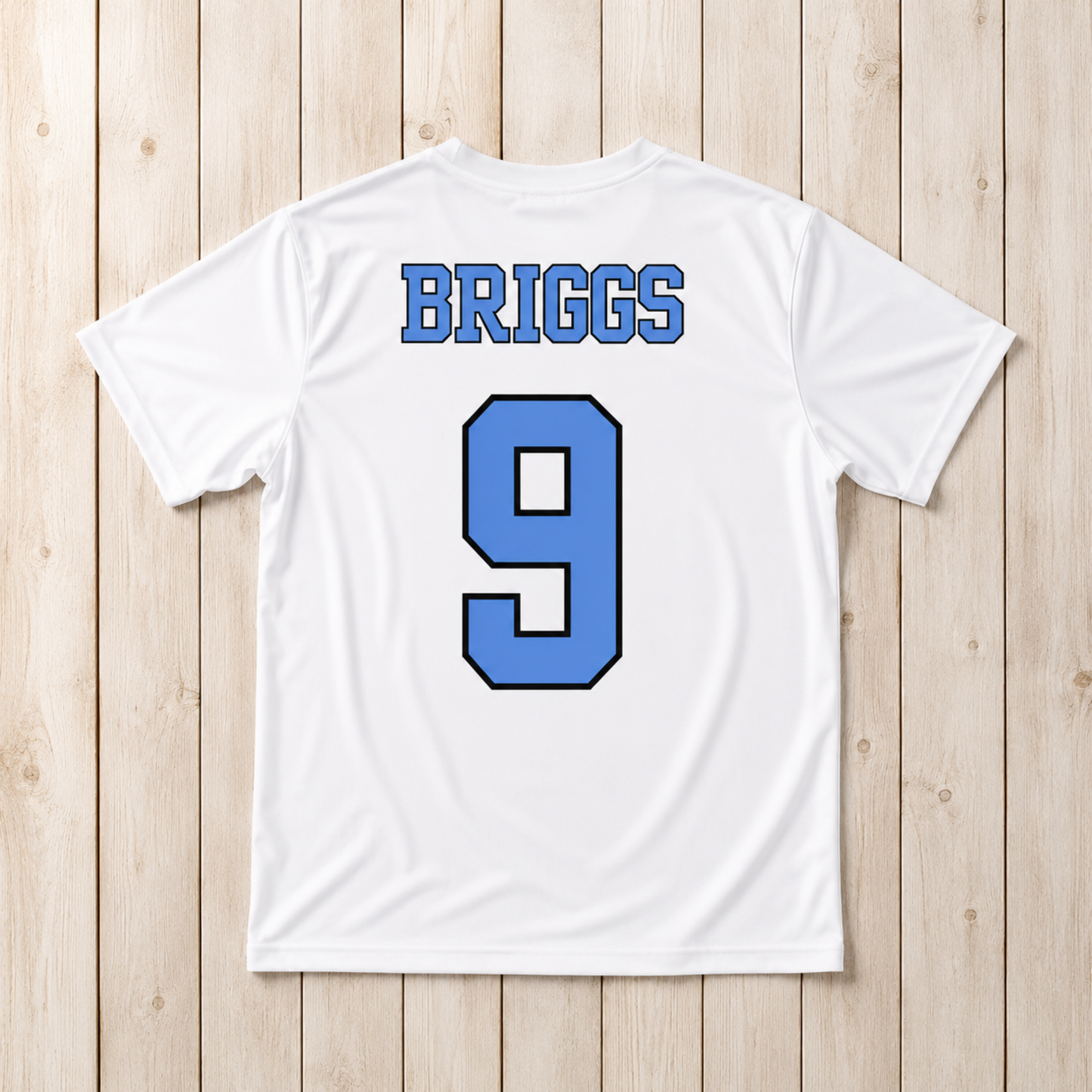 White short-sleeve athletic T-shirt laid flat on a light wood plank background, showing the back with the name BRIGGS in blue block letters and the number 9 in blue with a black outline.