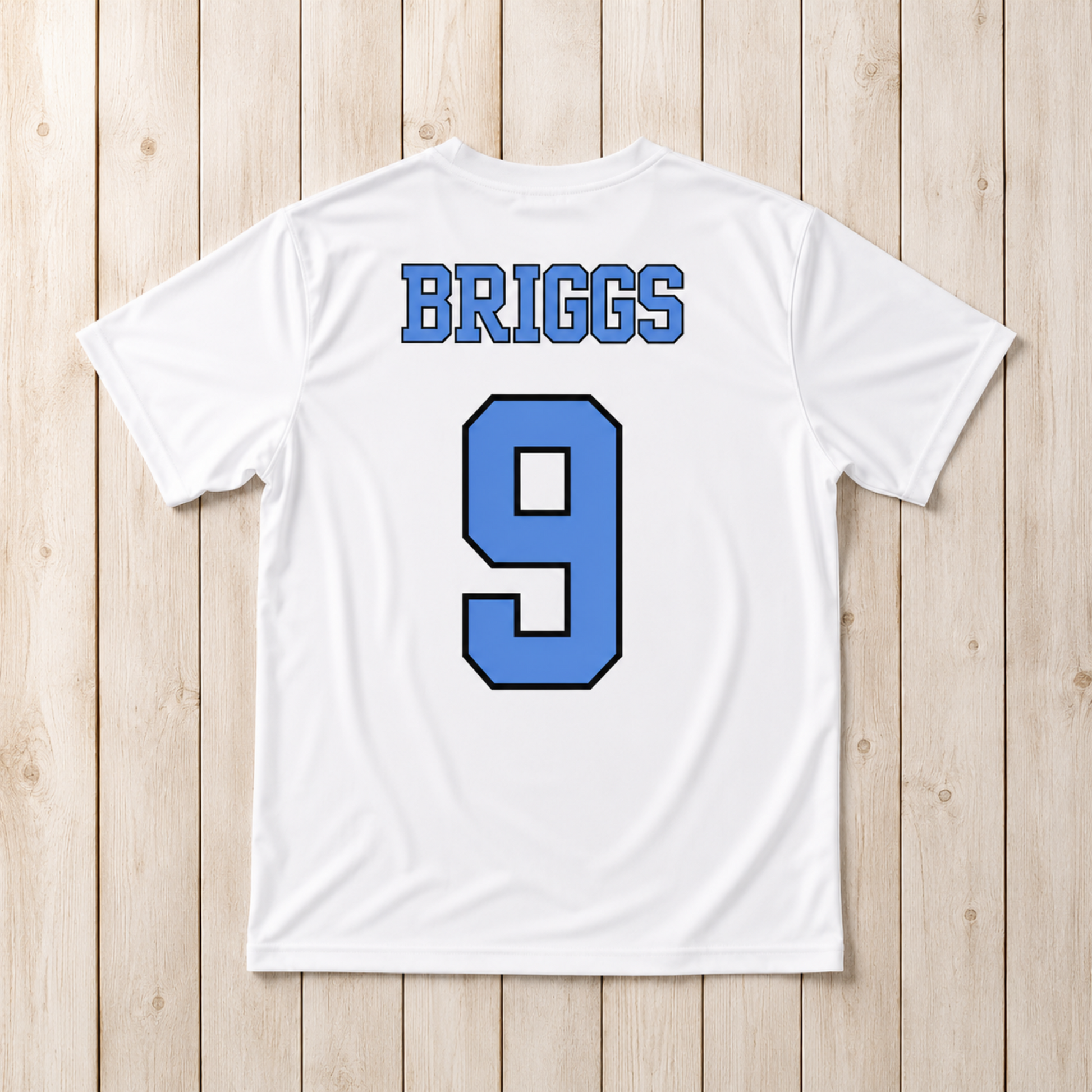 White short-sleeve athletic T-shirt laid flat on a light wood plank background, showing the back with the name BRIGGS in blue block letters and the number 9 in blue with a black outline.