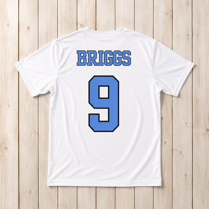 White short-sleeve athletic T-shirt laid flat on a light wood plank background, showing the back with the name BRIGGS in blue block letters and the number 9 in blue with a black outline.