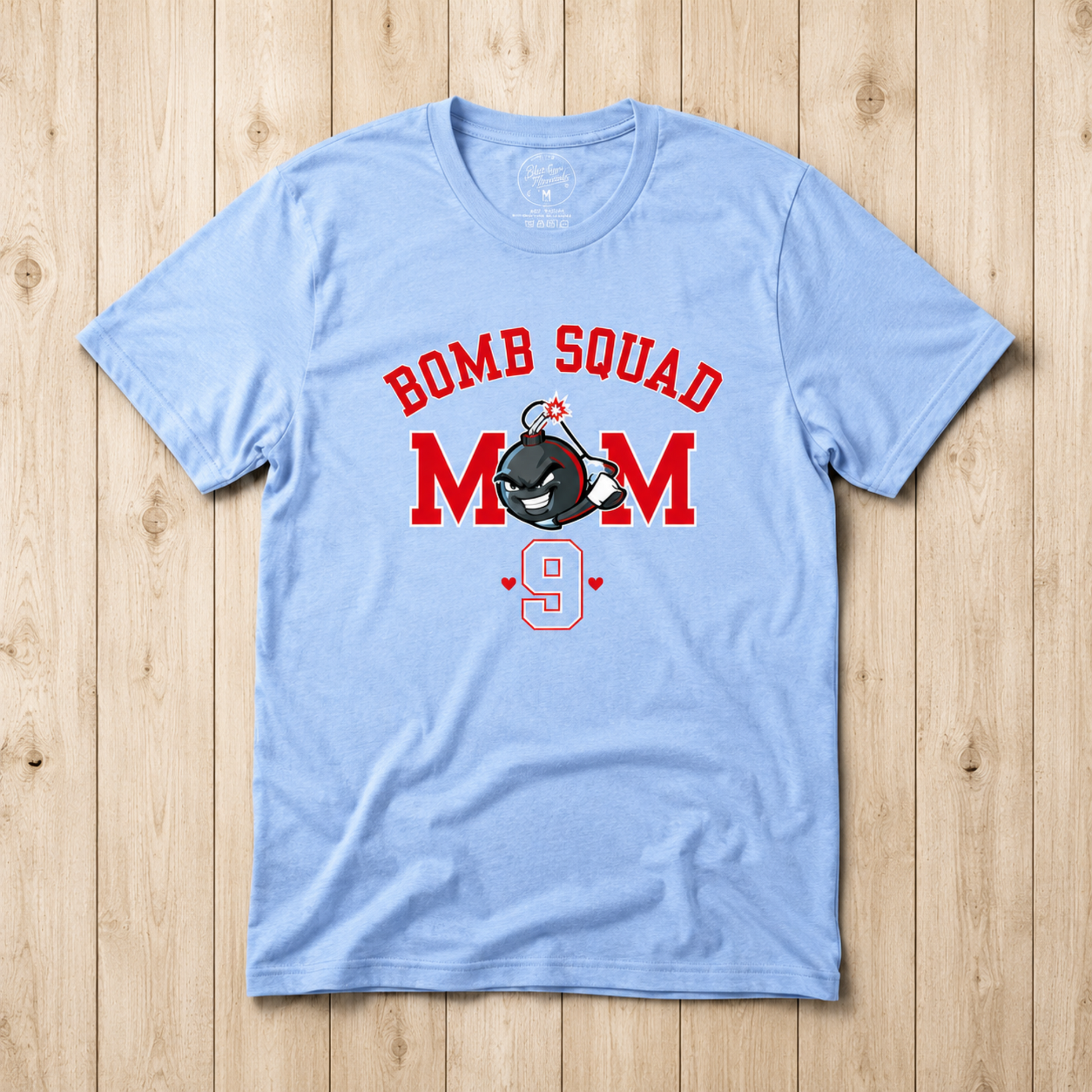 A light blue short-sleeve t-shirt with a 'BOMB SQUAD MOM 9' graphic, laid flat on a light wood plank background.