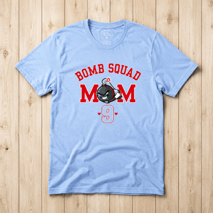 A light blue short-sleeve t-shirt with a 'BOMB SQUAD MOM 9' graphic, laid flat on a light wood plank background.