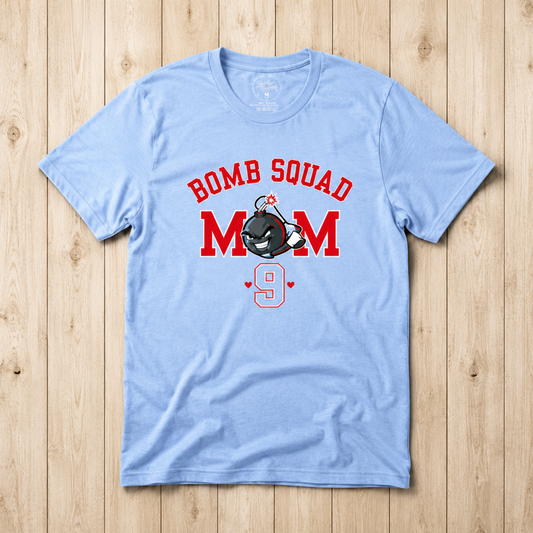 A light blue short-sleeve t-shirt with a 'BOMB SQUAD MOM 9' graphic, laid flat on a light wood plank background.