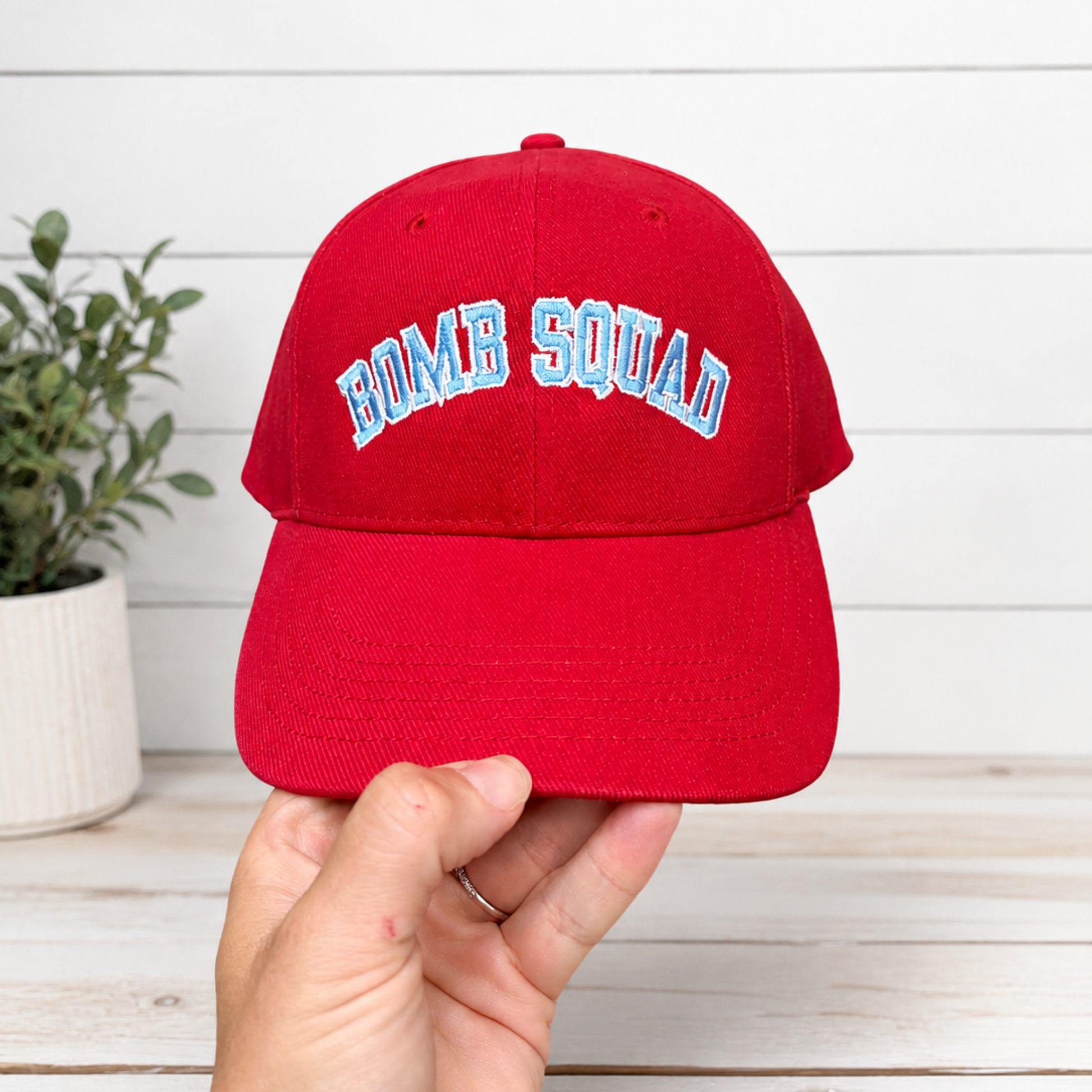 A red baseball cap with 'BOMB SQUAD' embroidered in white and light blue arched lettering, held by a hand against a white wood plank background with a small potted plant on the left.