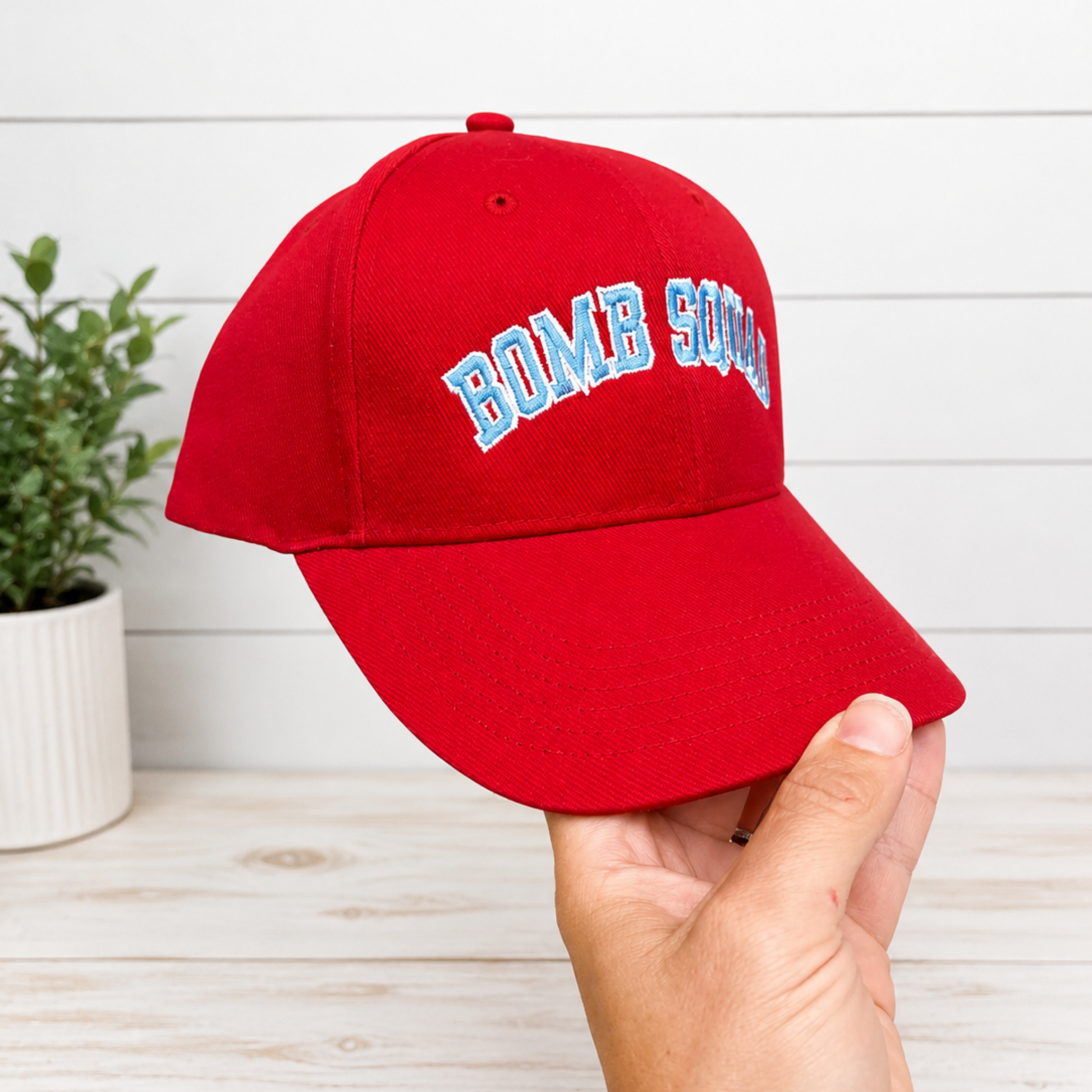 A red baseball cap with 'BOMB SQUAD' embroidered in white and light blue arched lettering, held by a hand against a white wood-paneled background.