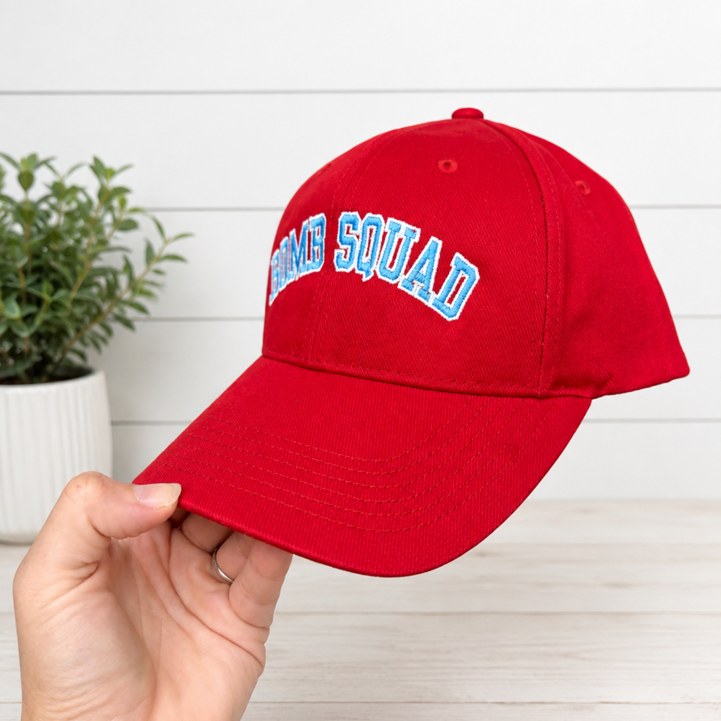 A red baseball cap with 'BOMB SQUAD' embroidered in light blue and white arched lettering on the front, held by a hand against a white wood-paneled background with a small potted plant.