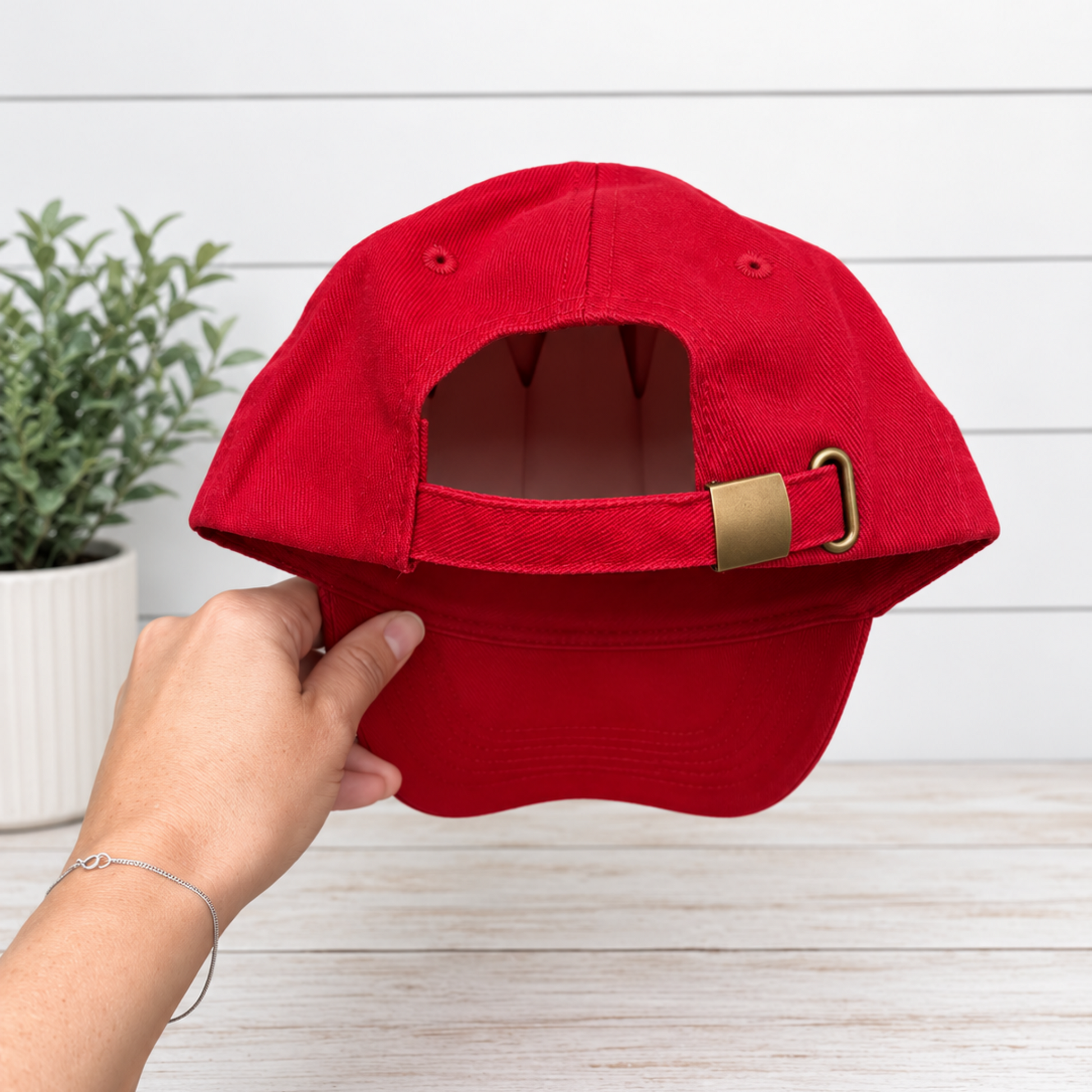 A red baseball cap held by a hand, showing the back strap and gold-tone buckle, set against a white wood-paneled wall and light wood tabletop.