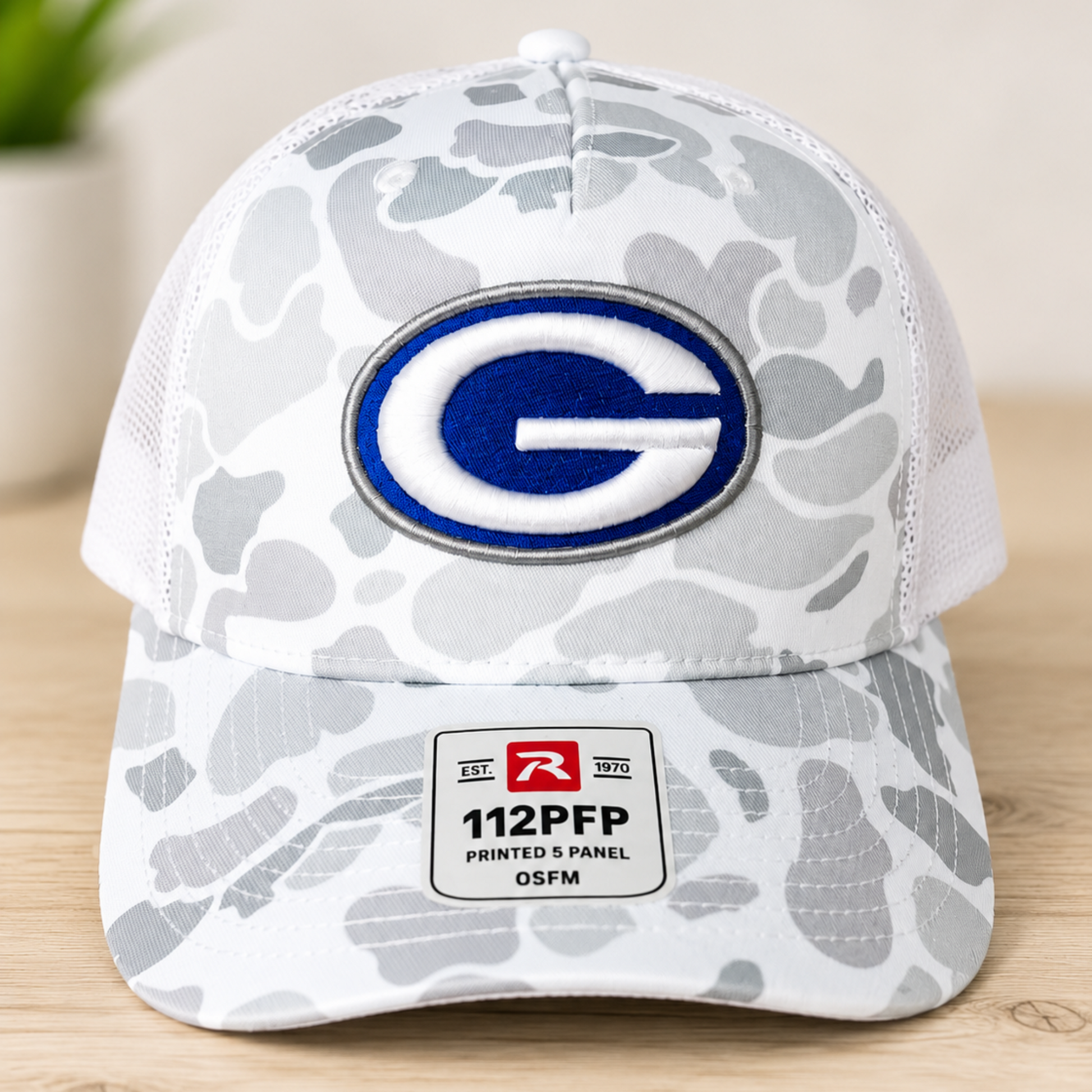 A white and gray camouflage trucker hat with a blue and white georgetown eagles logo patch on the front, resting on a light wood surface.