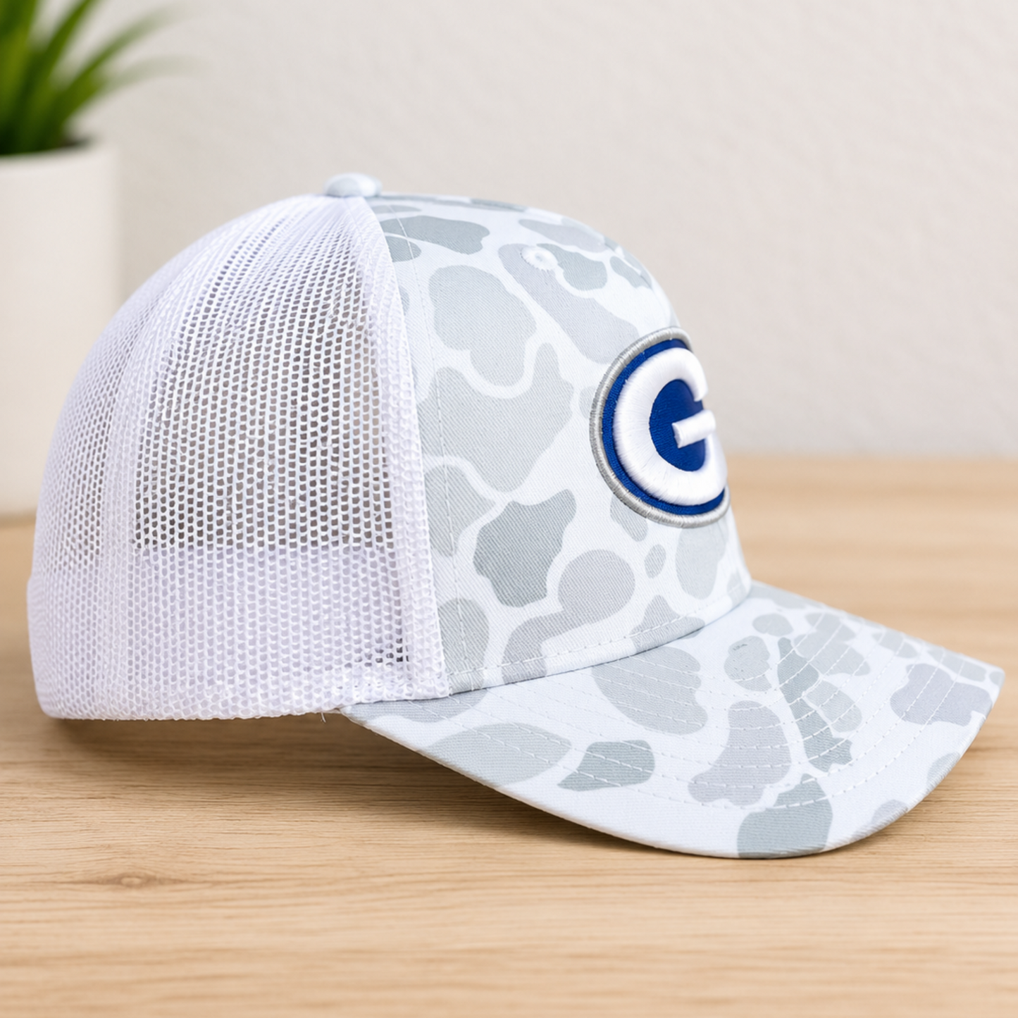 A white and gray camouflage trucker hat with a blue and white 'G' logo patch on the front, displayed on a light wood surface with a blurred indoor background.