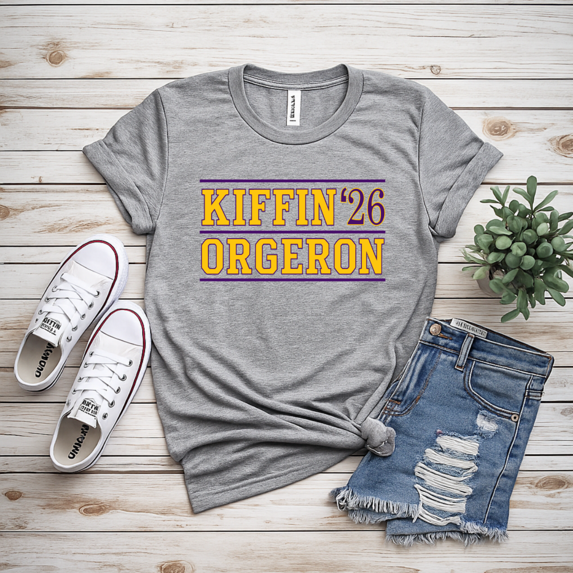 A gray heather graphic t-shirt with 'KIFFIN '26 ORGERON' printed in yellow and maroon, styled in a flat lay with distressed denim shorts, white sneakers, and a small succulent on a light wood plank background.