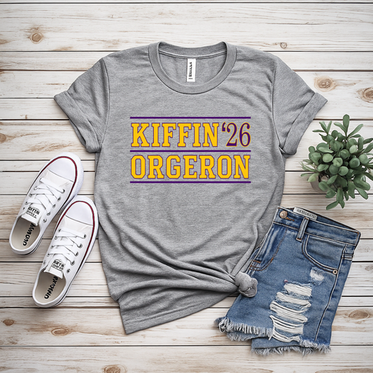A gray heather graphic t-shirt with 'KIFFIN '26 ORGERON' printed in yellow and maroon, styled in a flat lay with distressed denim shorts, white sneakers, and a small succulent on a light wood plank background.