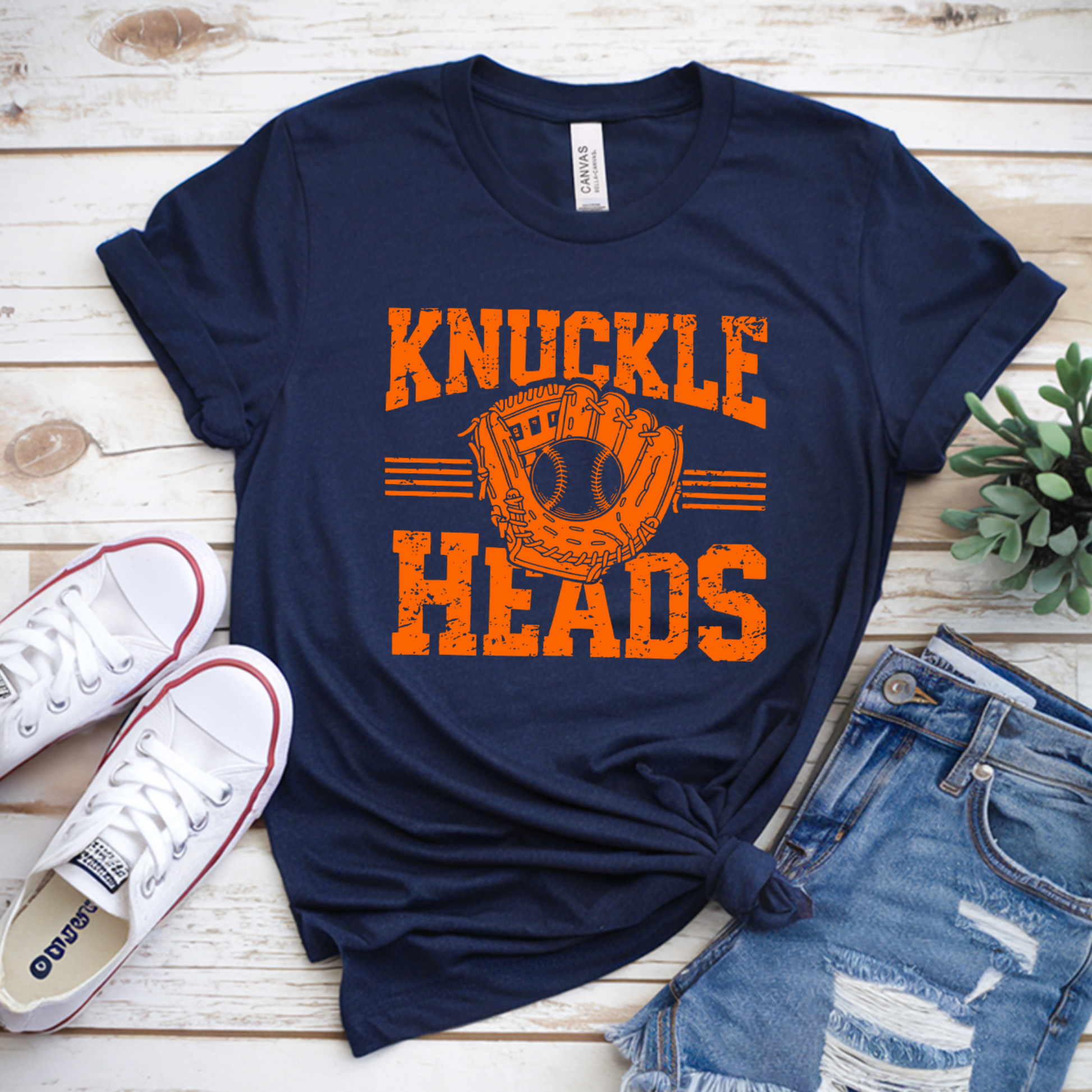 Navy t-shirt with 'Knuckle Heads' print on a wooden surface with sneakers and shorts.