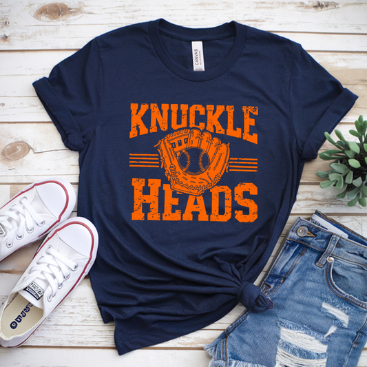 Navy t-shirt with 'Knuckle Heads' print on a wooden surface with sneakers and shorts.