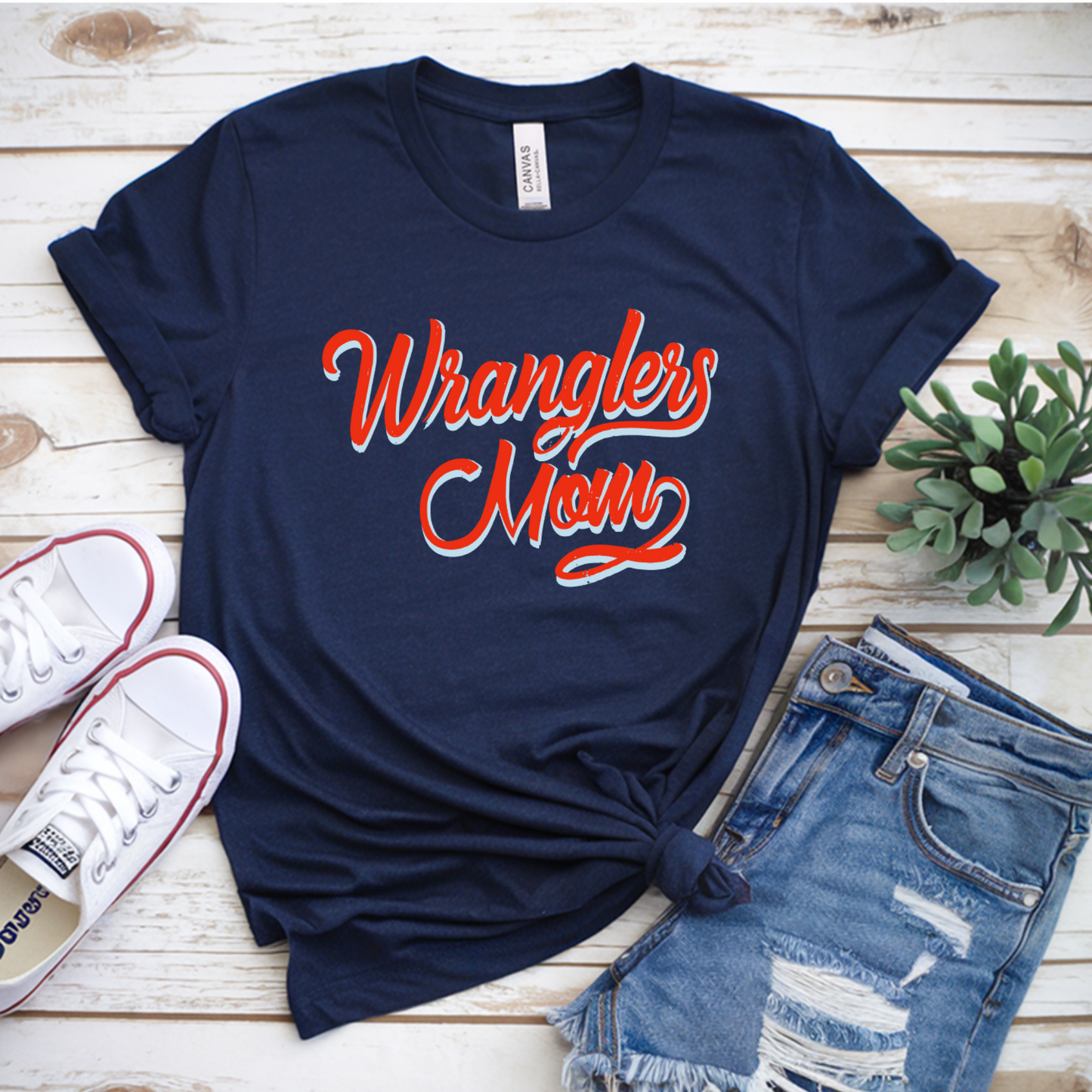 A navy blue graphic t-shirt with 'Wranglers Mom' printed in red and white script, styled in a flat lay with distressed denim shorts, white sneakers, and a small potted plant on a light wood background.