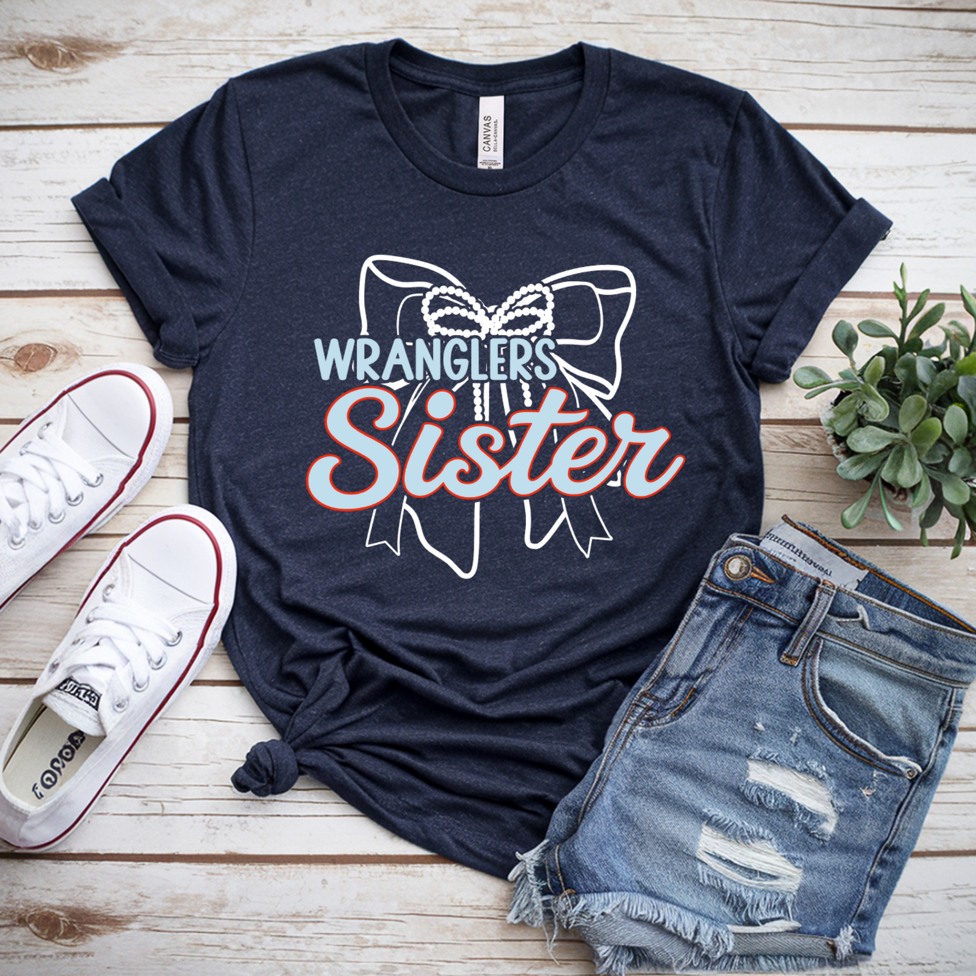 A navy blue graphic t-shirt featuring the text 'WRANGLERS Sister' with a bow illustration, styled in a flat lay on a light wood background with white sneakers and distressed denim shorts.
