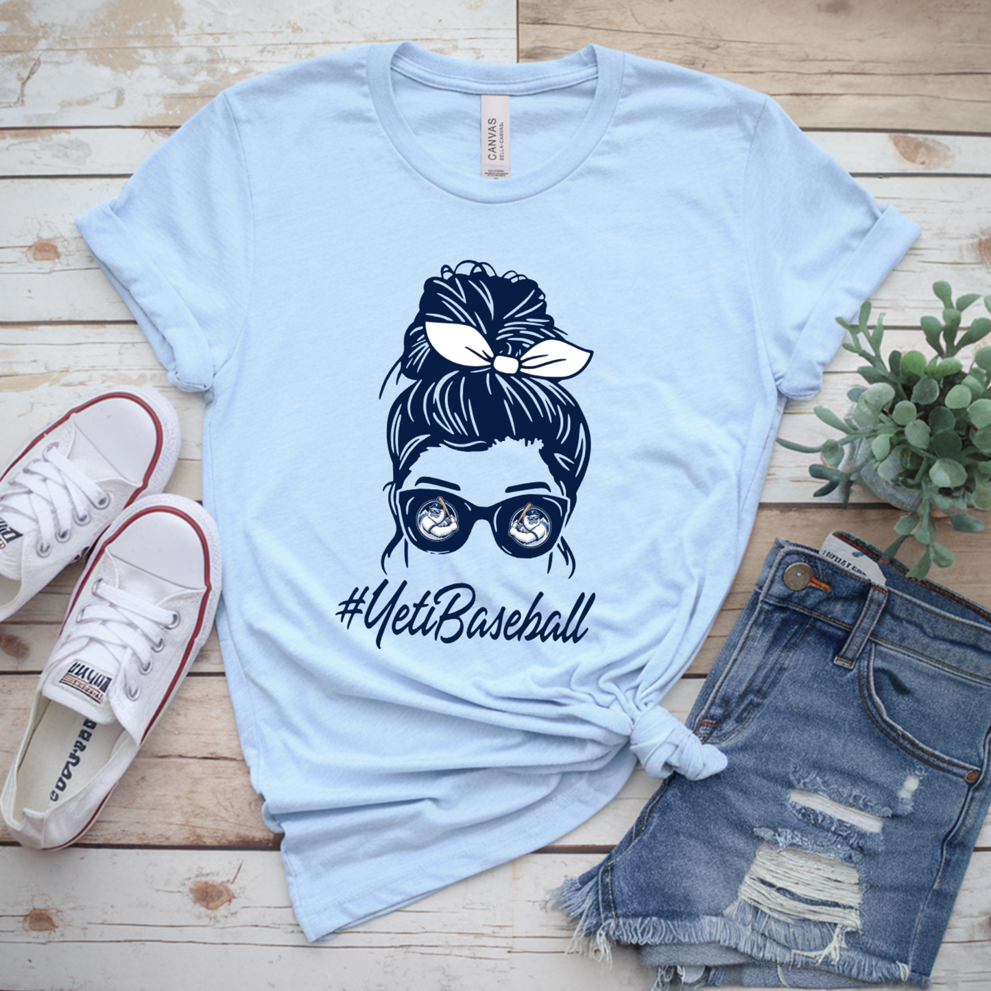Light blue t-shirt with graphic design and text, paired with denim shorts and sneakers on a wooden surface.