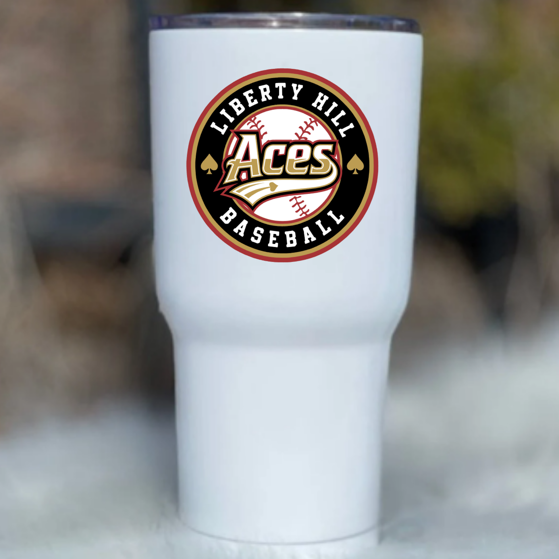A white insulated tumbler with a clear lid, featuring a circular 'Liberty Hill Aces Baseball' logo printed on the front, set against a blurred outdoor background.