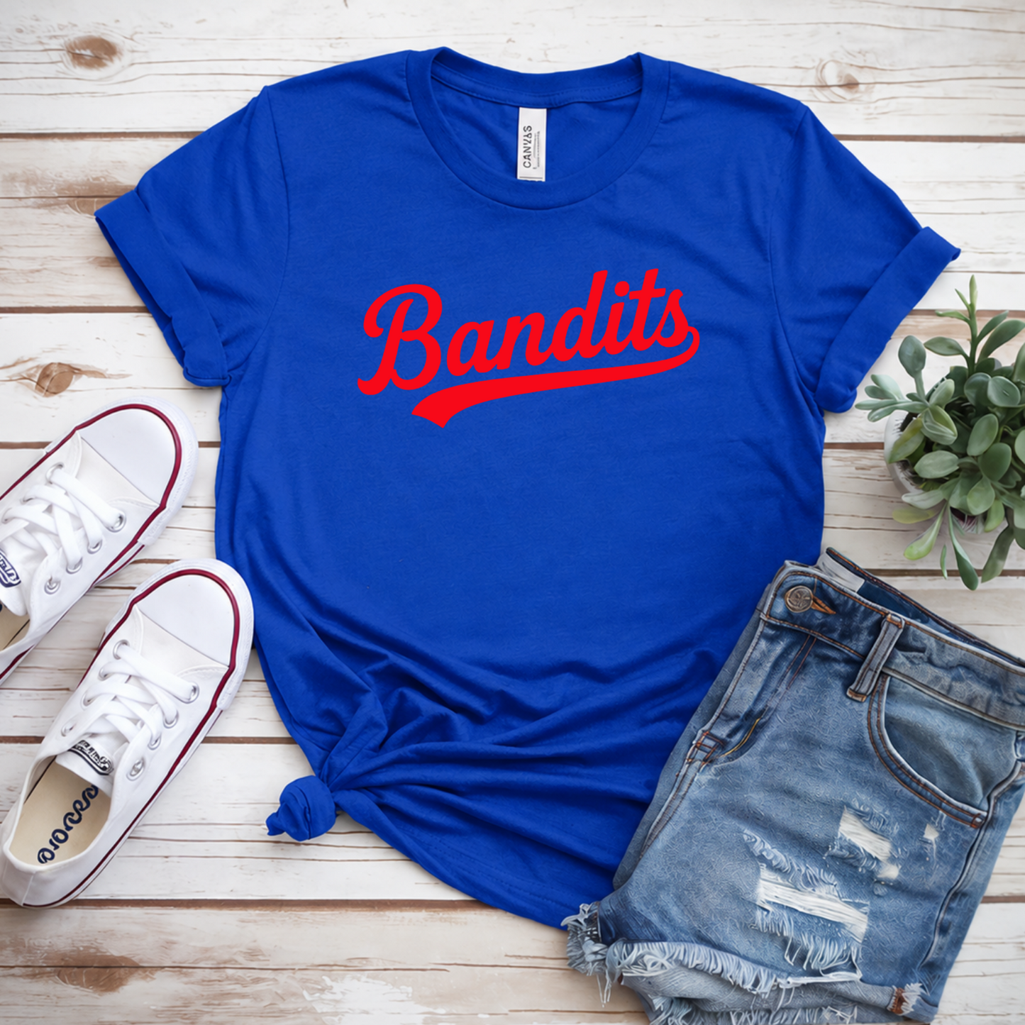 Blue t-shirt with 'Bandits' text, white sneakers, denim shorts, and a plant on a wooden surface.